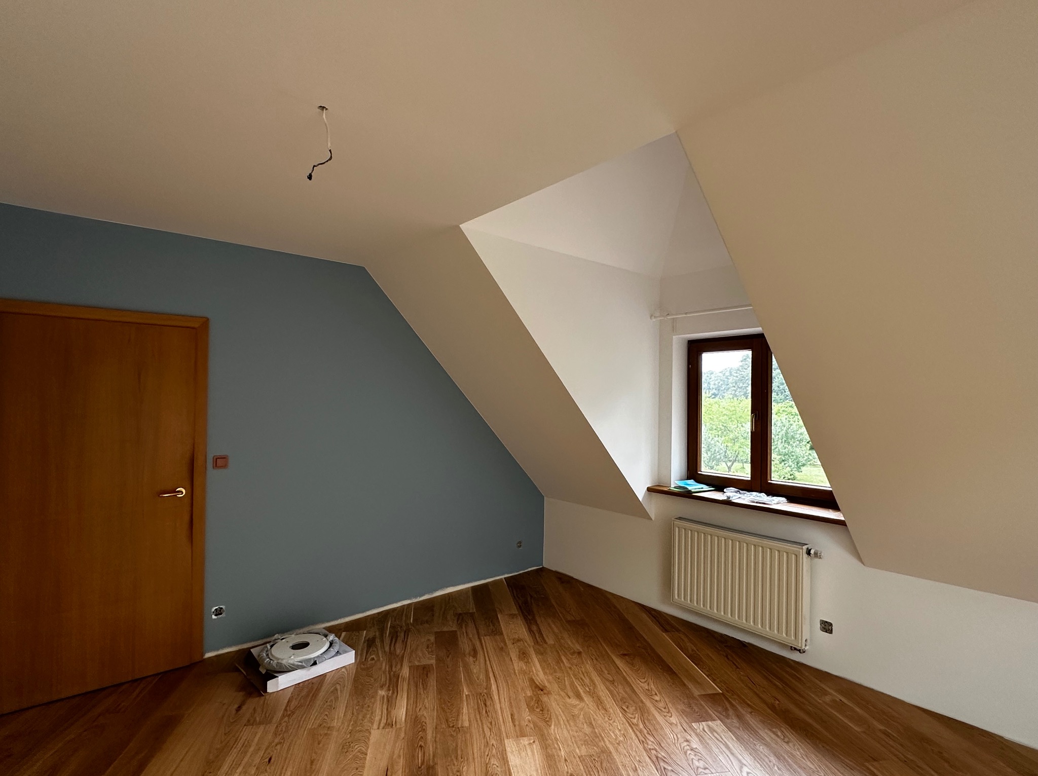 Pokój na poddaszu po malowaniu: błękitna ściana kontrastuje z bielą skosów, drewniana podłoga i okno z widokiem na zieleń. Widoczny grzejnik i elementy instalacji elektrycznej.