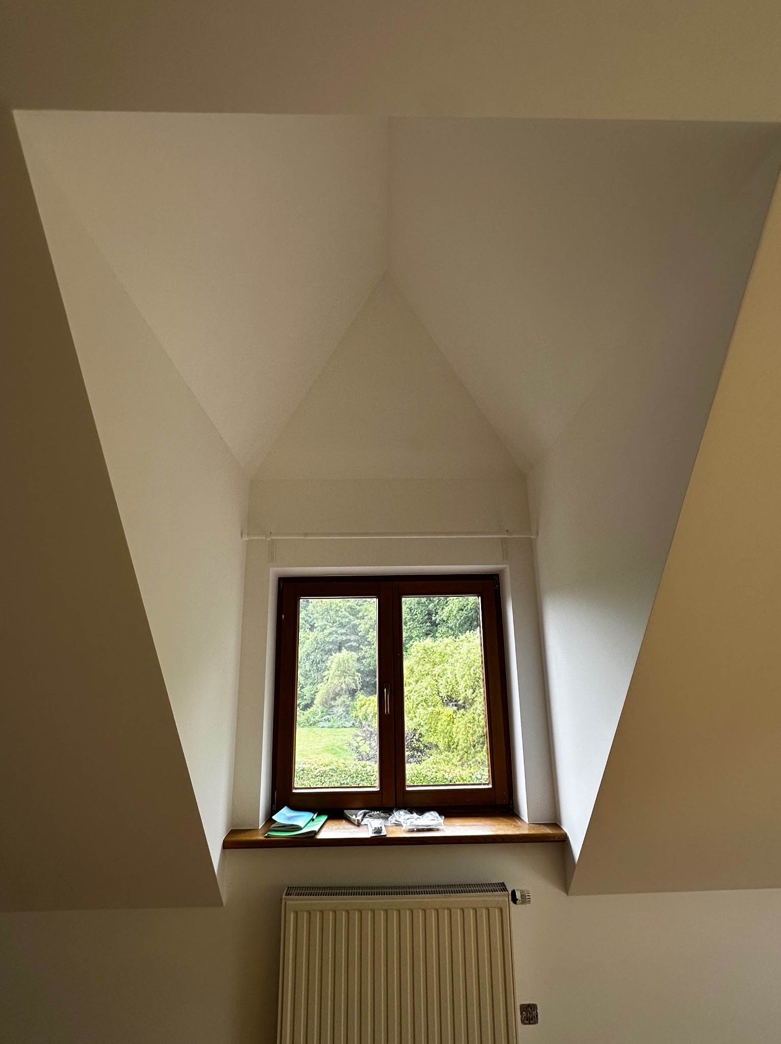 Poddasze po remoncie: świeżo pomalowane ściany w odcieniach bieli, drewniane okno z widokiem na zieleń i grzejnik. Skośne ściany tworzą unikalny kształt pomieszczenia.