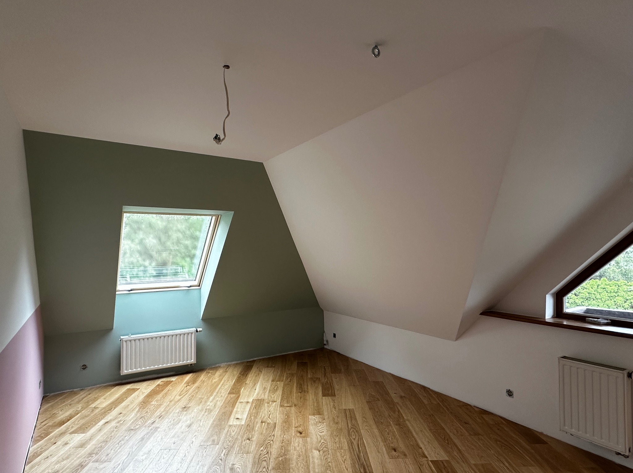 Pokój na poddaszu po malowaniu ścian w trzech kolorach: białym, zielonym i różowym. Widoczne okna dachowe i drewniana podłoga. Częściowo widoczny grzejnik.