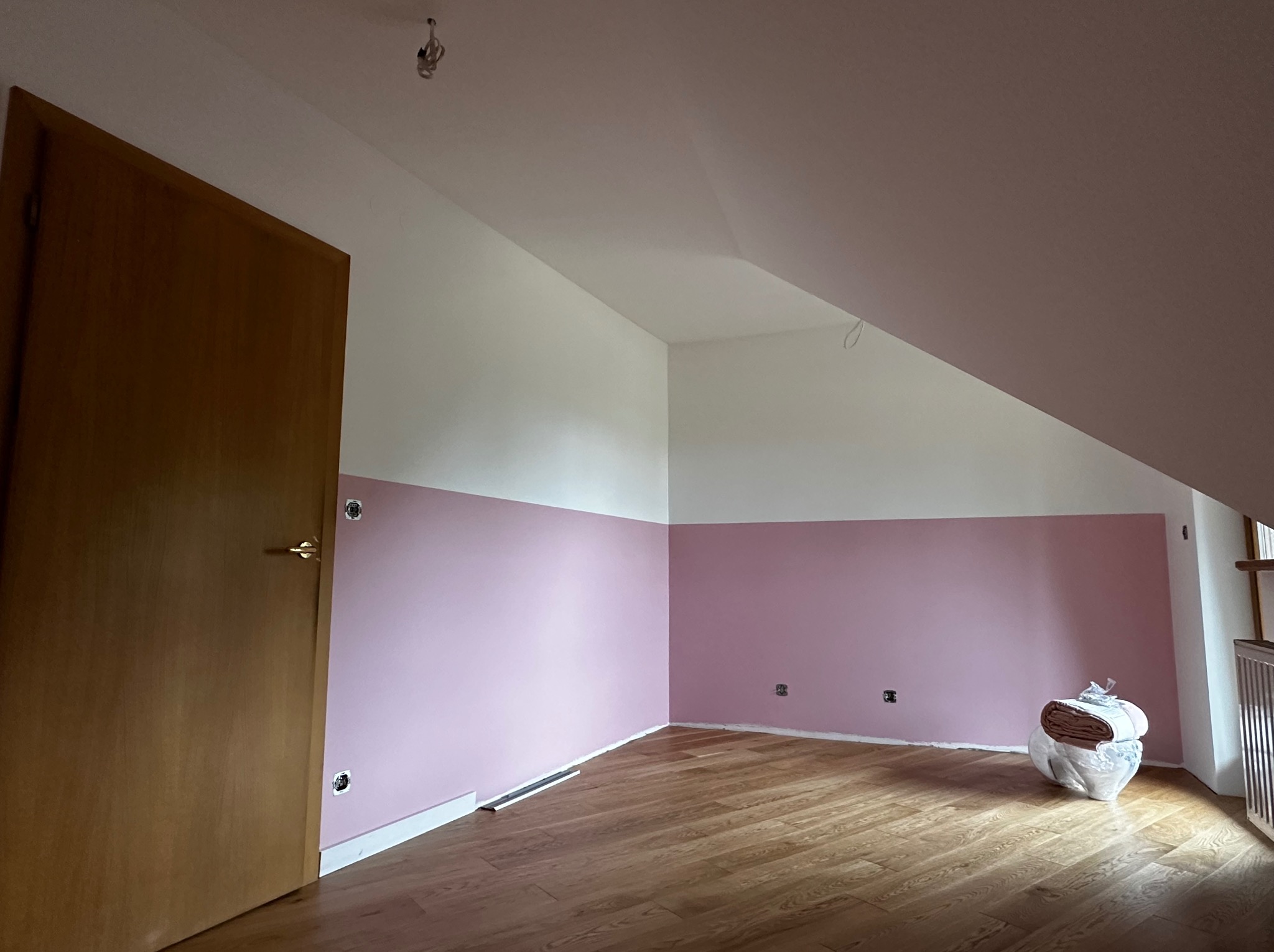 Pokój na poddaszu po malowaniu: różowe ściany do połowy wysokości, biała góra. Drewniana podłoga, brązowe drzwi po lewej. Widoczne gniazdka elektryczne i pakunek materiałów.