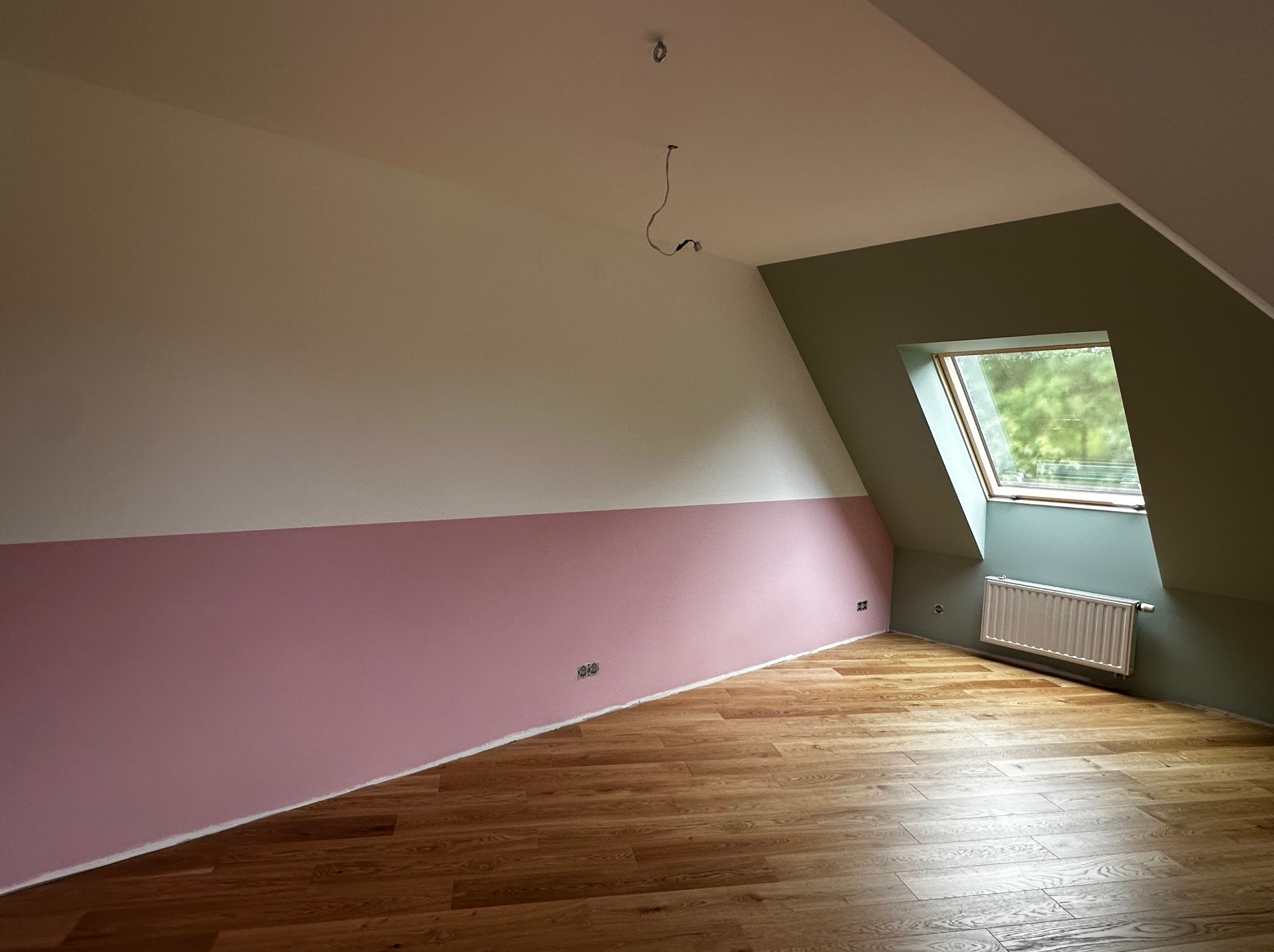 Pokój na poddaszu po remoncie: drewniana podłoga, ściany pomalowane na biało, różowo i zielono. Widoczne okno dachowe i grzejnik. W suficie wystają kable.