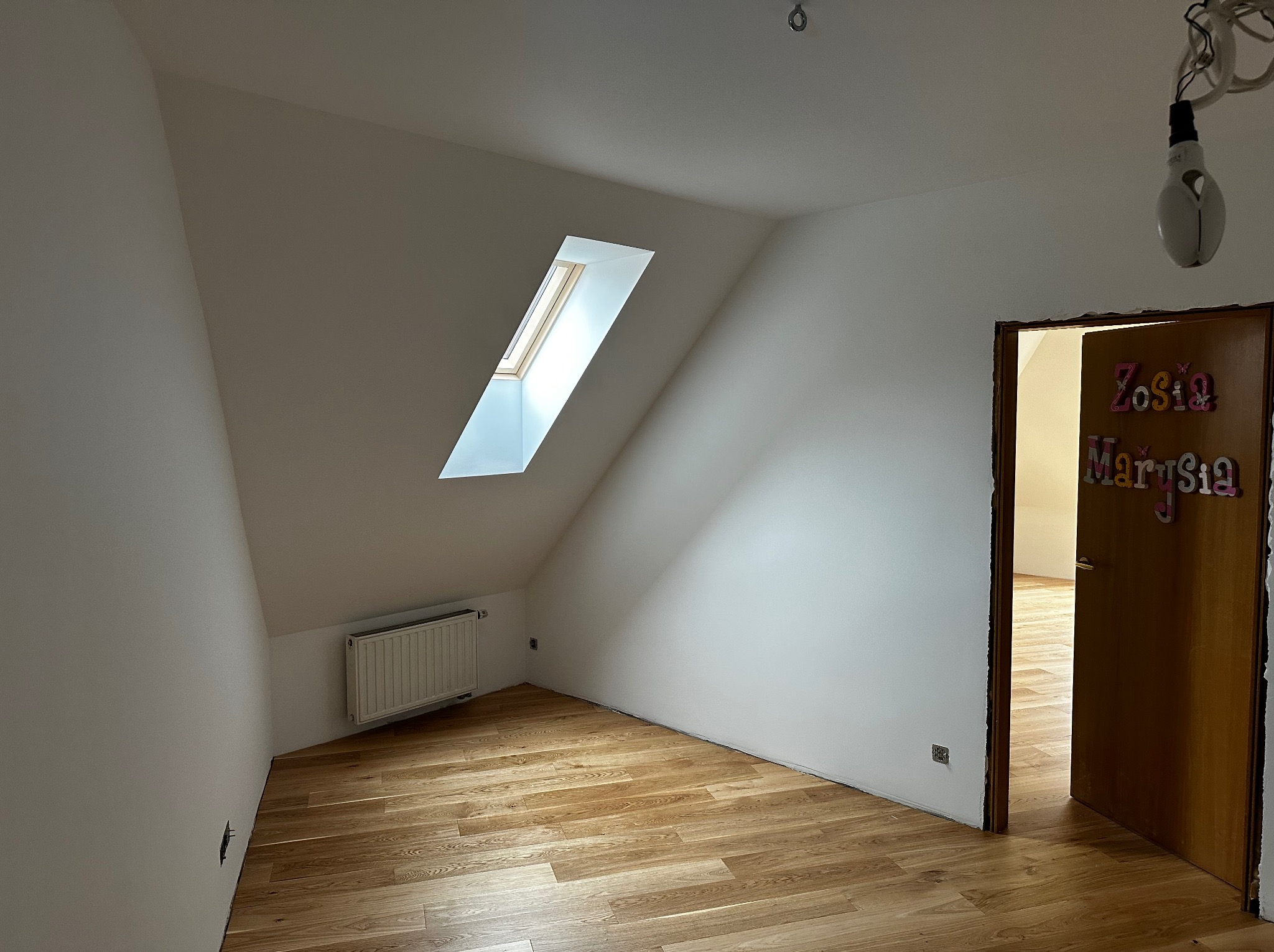 Jasne poddasze z oknem dachowym, świeżo pomalowane ściany na biało, drewniana podłoga. Widoczny grzejnik i fragment drzwi z dekoracjami.