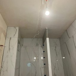 Bohdan Ostradchuk - Łazienka w trakcie remontu: białe płytki imitujące marmur na ścianach, surowy sufit z widocznymi przewodami i żarówką. Widoczne narzędzia do układania płytek.