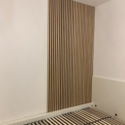 Bohdan Ostradchuk - Lamelki drewniane na ścianie w sypialni, nad ramą łóżka bez materaca. Pionowe panele dekoracyjne w jasnym odcieniu drewna, minimalistyczny design.