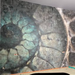 Bohdan Ostradchuk - Ściana z unikalną, wzorzystą tapetą w odcieniach szarości i turkusu, z widocznymi gniazdkami elektrycznymi. Podłoga w trakcie prac wykończeniowych.