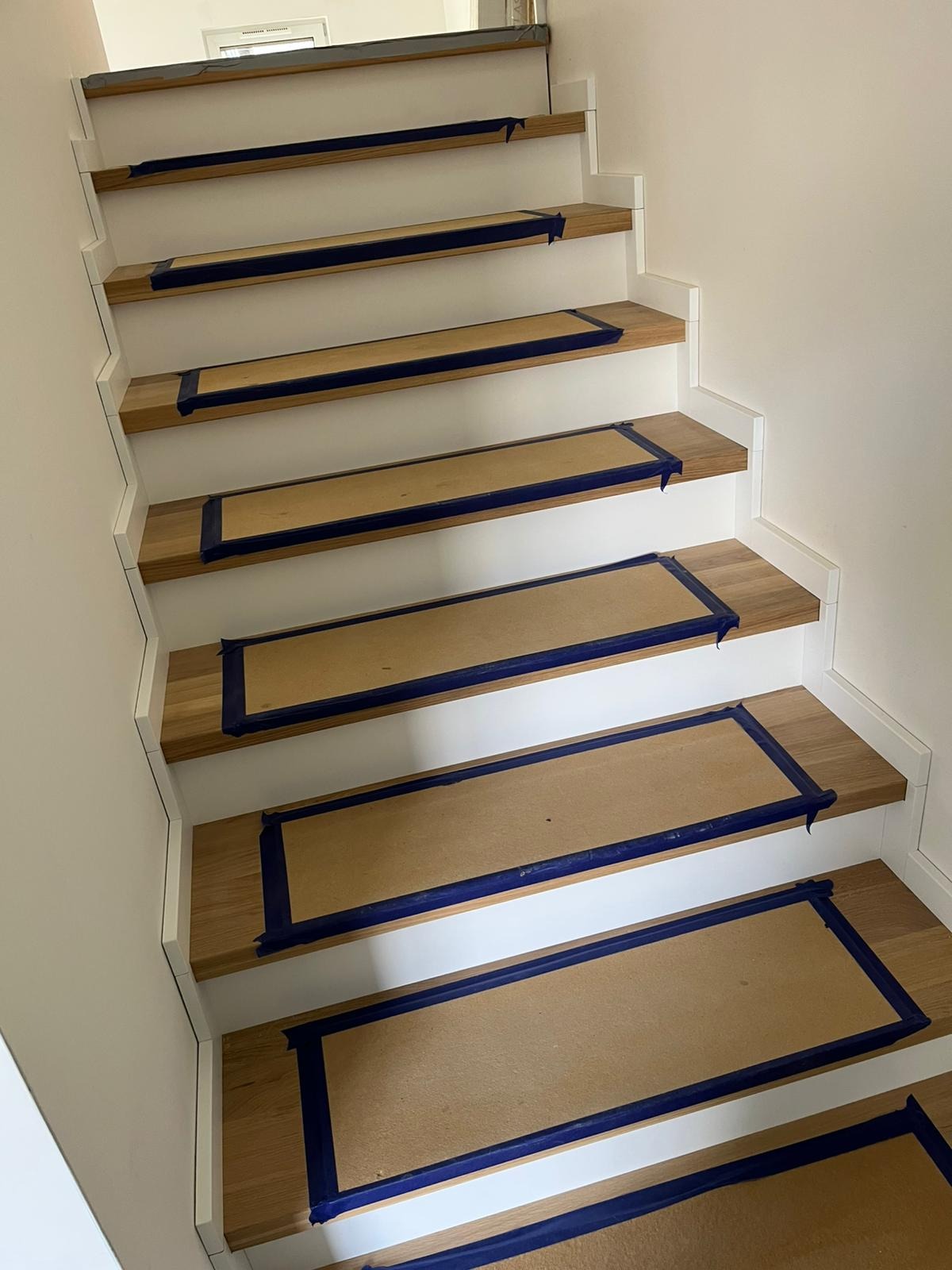 Schody drewniane z białymi podstopnicami zabezpieczone kartonem i niebieską taśmą malarską przed dalszymi pracami wykończeniowymi, widok z dołu.
