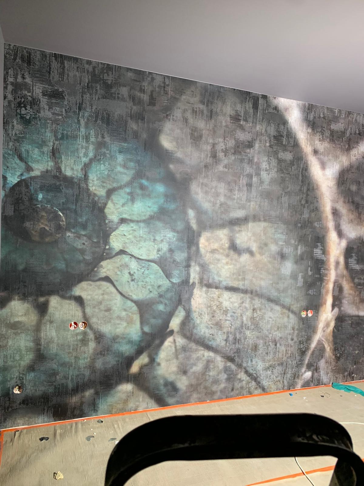 Ściana z unikalną, wzorzystą tapetą w odcieniach szarości i turkusu, z widocznymi gniazdkami elektrycznymi. Podłoga w trakcie prac wykończeniowych.