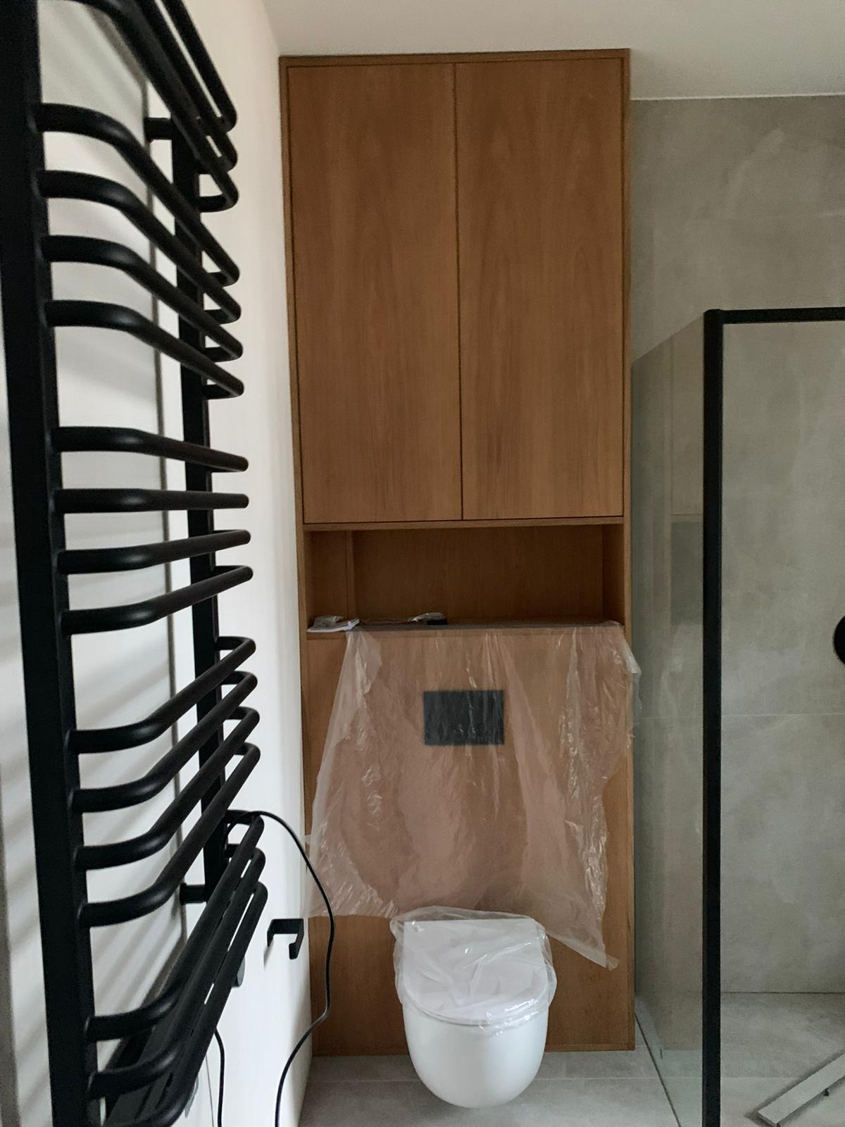 Łazienka po remoncie w Warszawie: zabudowa z szafkami z jasnego drewna, wisząca toaleta w folii ochronnej i czarny grzejnik drabinkowy.