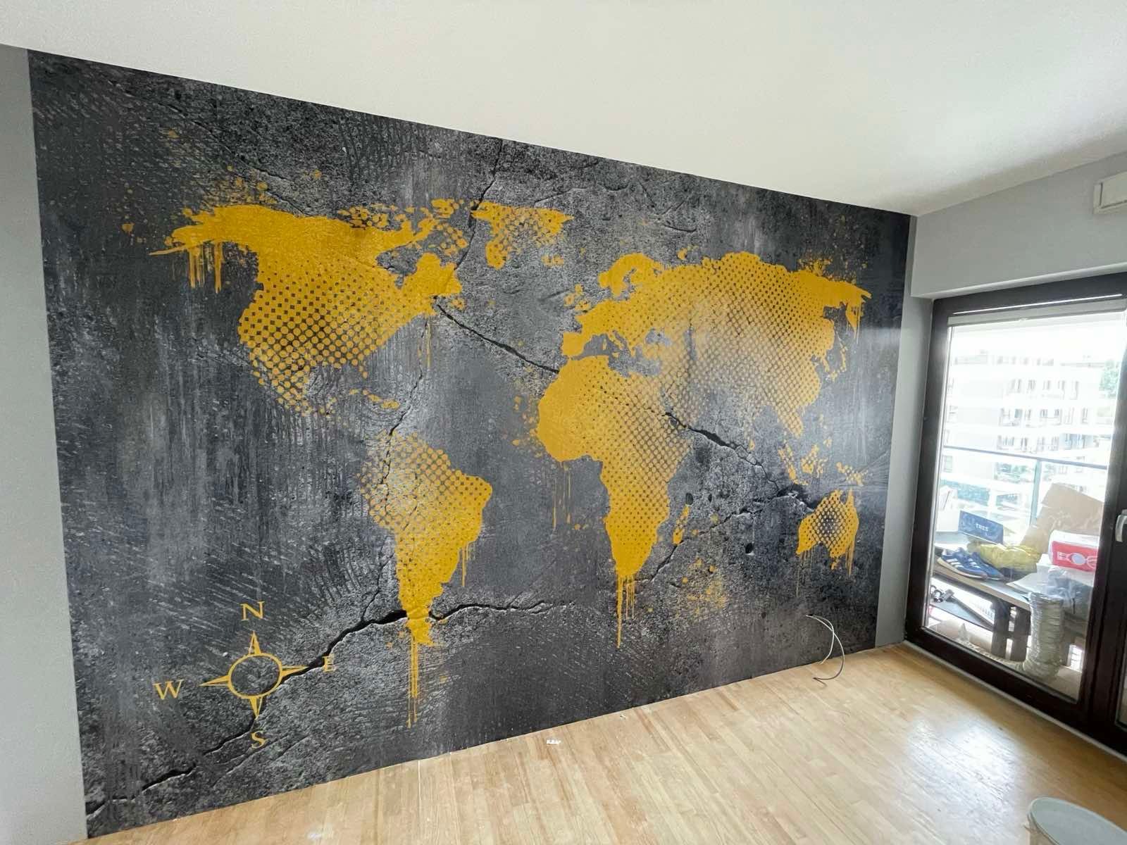 Ściana z mapą świata w odcieniach szarości i złota, stylizowana na popękaną powierzchnię. Widoczny kompas w lewym dolnym rogu i drewniana podłoga.