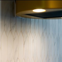 COMEX SPÓŁKA Z OGRANICZONĄ ODPOWIEDZIALNOŚCIĄ - Fragment ściany z marmurowymi płytkami ułożonymi w geometryczny wzór, oświetlonymi przez lampy okrągłego okapu kuchennego w kolorze złota.