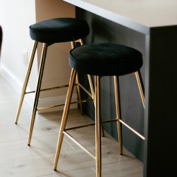 COMEX SPÓŁKA Z OGRANICZONĄ ODPOWIEDZIALNOŚCIĄ - Dwa welurowe hokery z pozłacanymi nogami przy ciemnoszarym blacie kuchennym, na którym leżą ciastka na szklanych talerzykach.