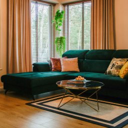 COMEX SPÓŁKA Z OGRANICZONĄ ODPOWIEDZIALNOŚCIĄ - Salon z zielonym narożnikiem, okrągłym stolikiem kawowym na metalowych nóżkach, drewnianą podłogą i zasłonami w odcieniach beżu. Na ścianie wisi obraz w czarnej ramie.