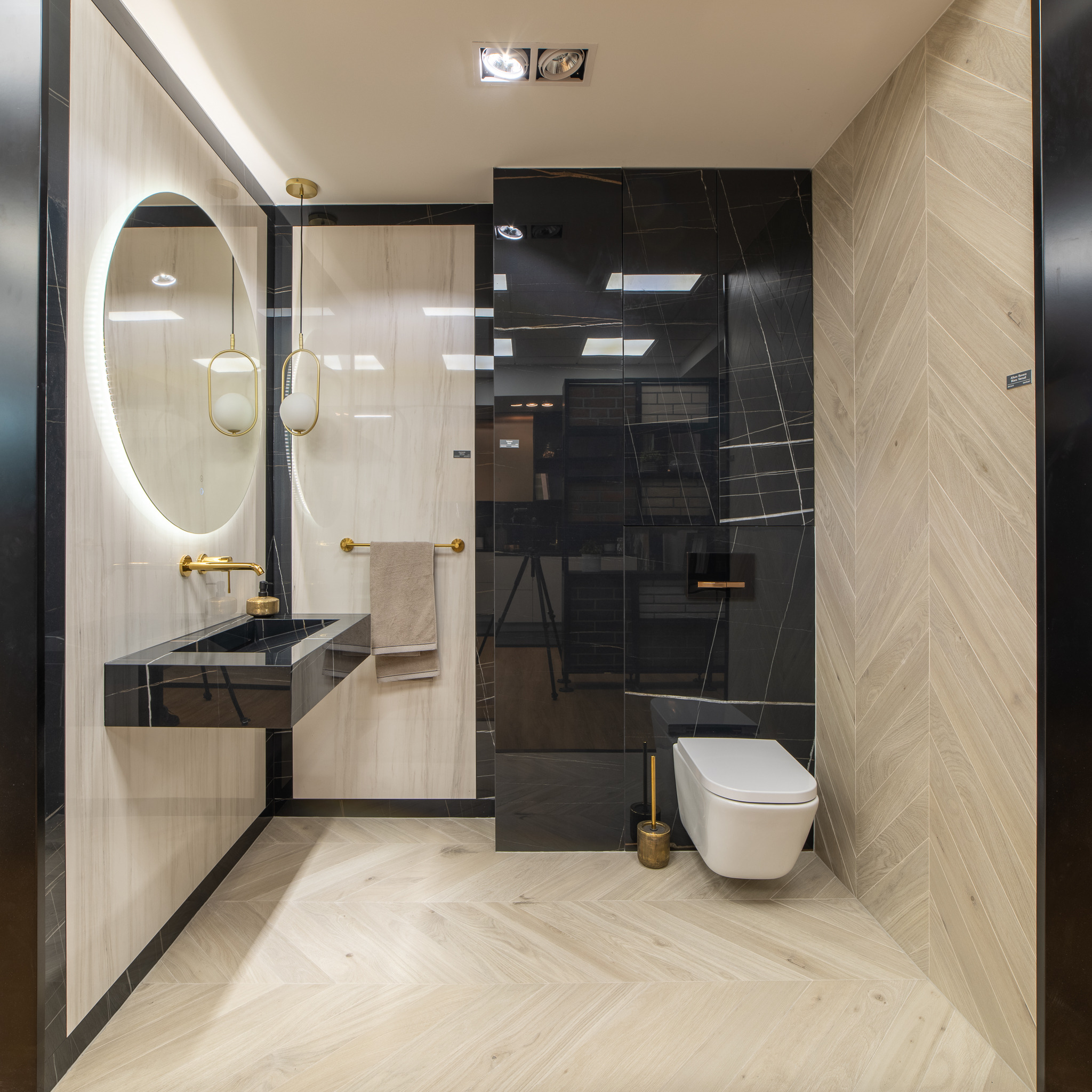 Nowoczesna łazienka z czarną umywalką na postumencie, okrągłym lustrem z podświetleniem LED, złotą armaturą i toaletą wiszącą.