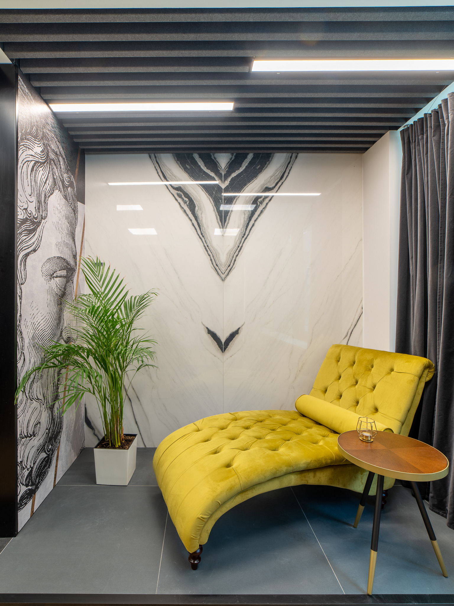 Żółty, pikowany szezlong z poduszką i małym stolikiem w minimalistycznym wnętrzu z marmurową ścianą i motywem graficznym twarzy na ścianie obok rośliny doniczkowej.