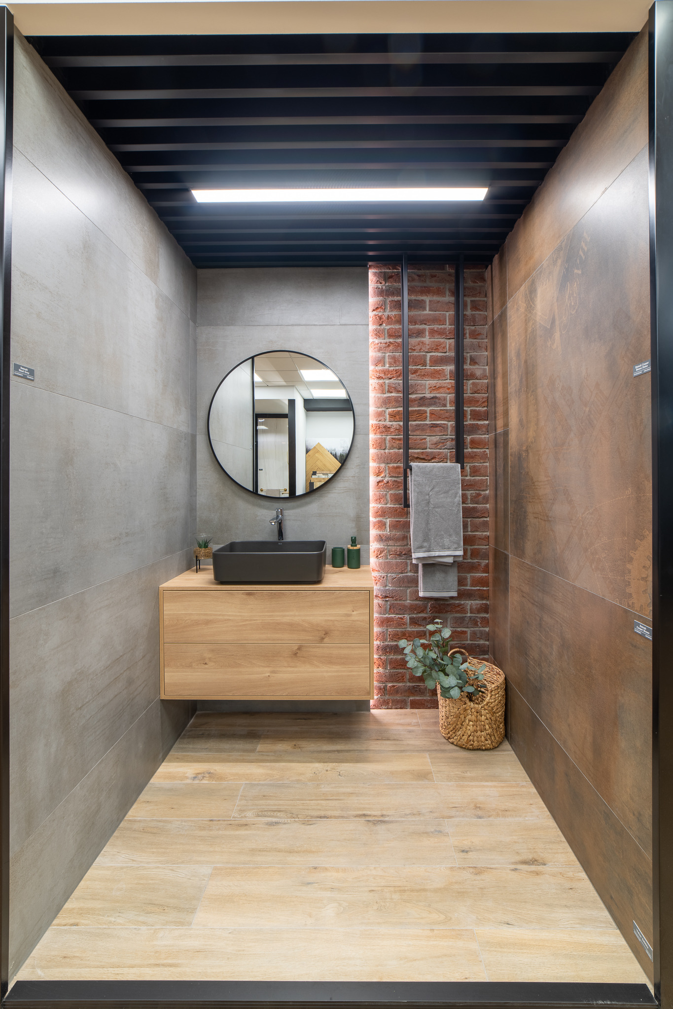 Nowoczesna łazienka z elementami cegły, umywalką nablatową na drewnianej szafce, okrągłym lustrem i ręcznikami na metalowym wieszaku.