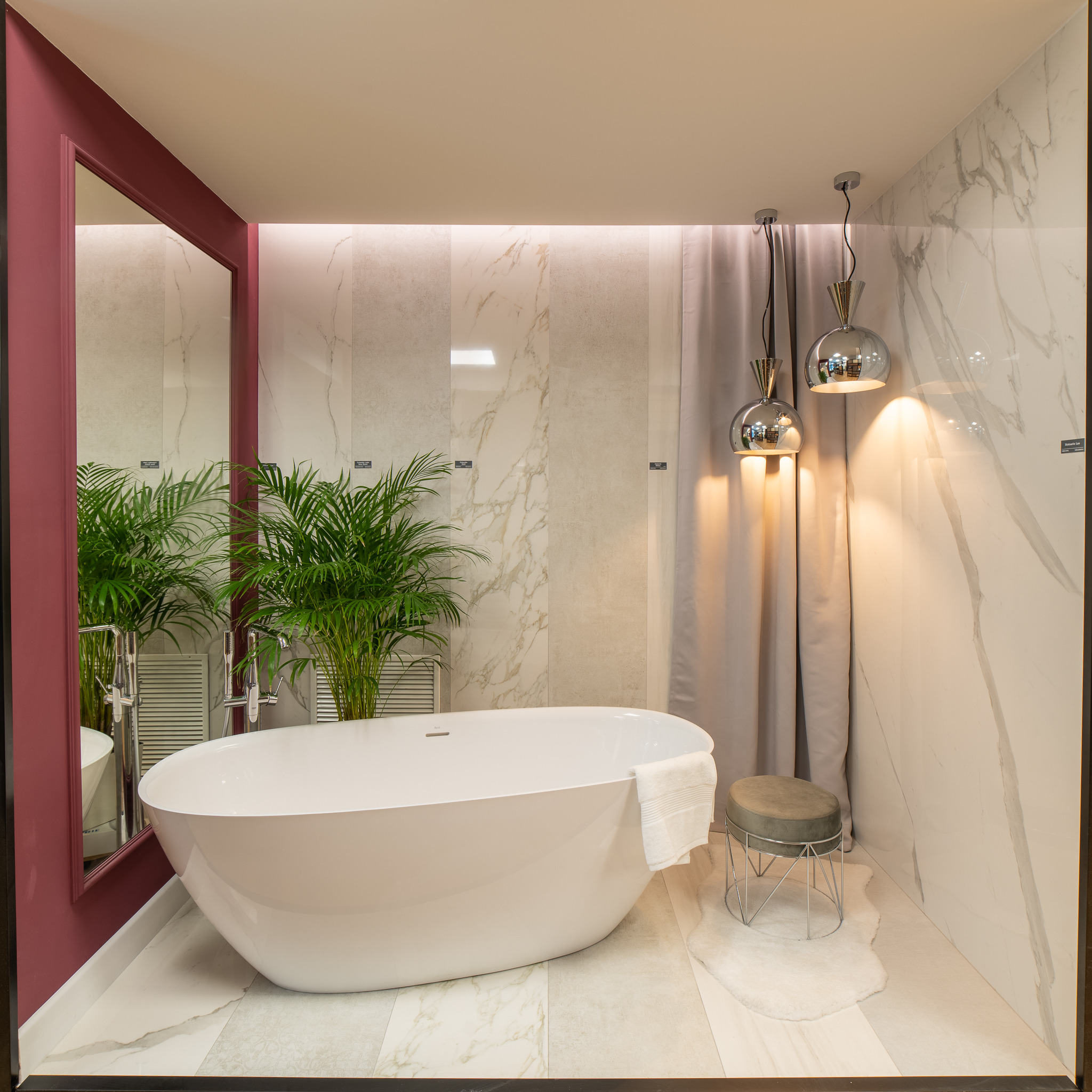 Nowoczesna łazienka z wolnostojącą białą wanną, marmurowymi płytkami ściennymi, lustrem w bordowej ramie, rośliną doniczkową i chromowanymi lampami wiszącymi.