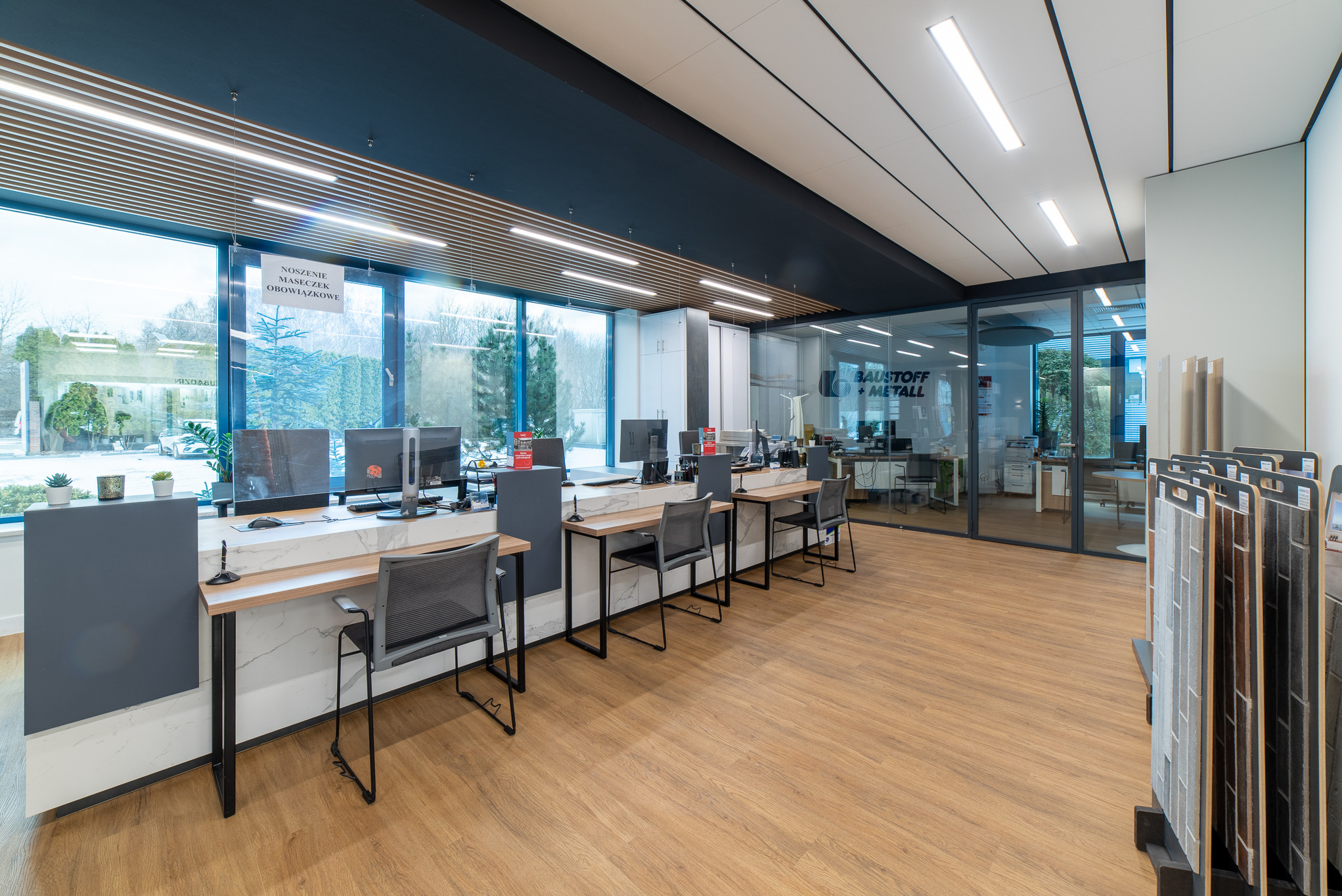 Nowoczesne biuro z drewnianą podłogą, stanowiskami pracy wzdłuż okna i ekspozycją próbek materiałów wykończeniowych.