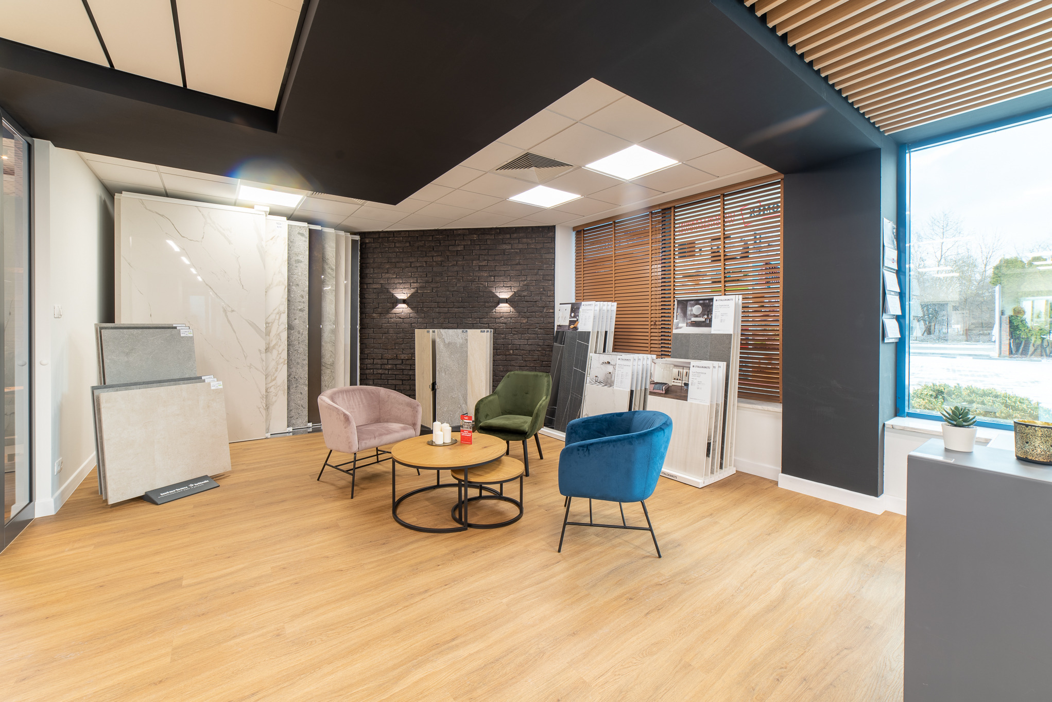 Salon z ekspozycją płytek ceramicznych i drewnopodobną podłogą, z trzema welurowymi krzesłami w odcieniach różu, zieleni i błękitu oraz okrągłymi stolikami kawowymi.