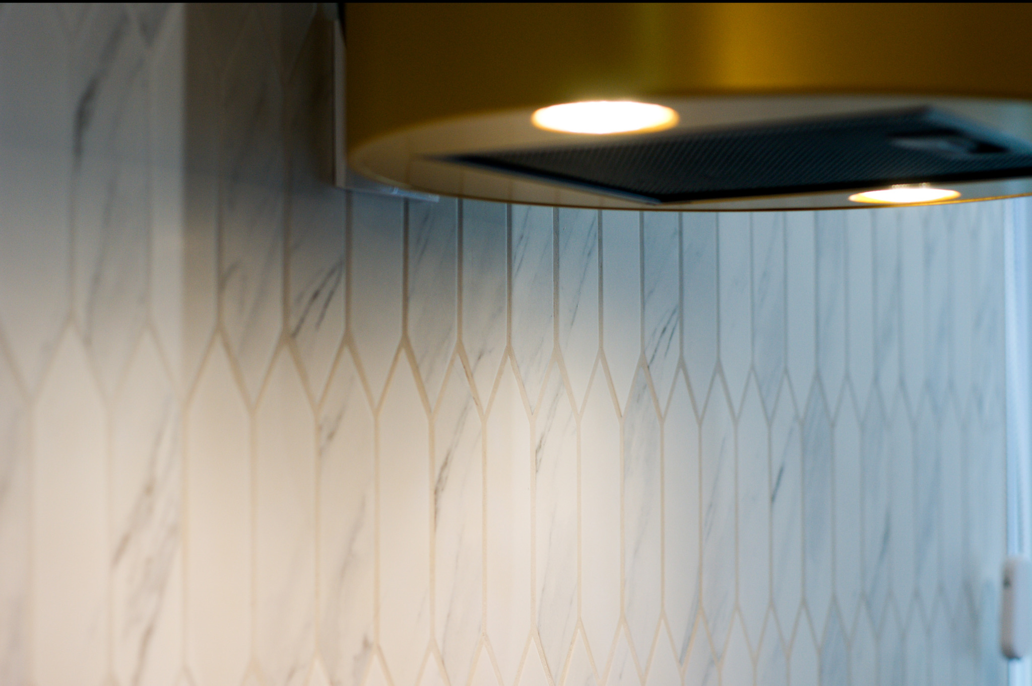 Fragment ściany z marmurowymi płytkami ułożonymi w geometryczny wzór, oświetlonymi przez lampy okrągłego okapu kuchennego w kolorze złota.