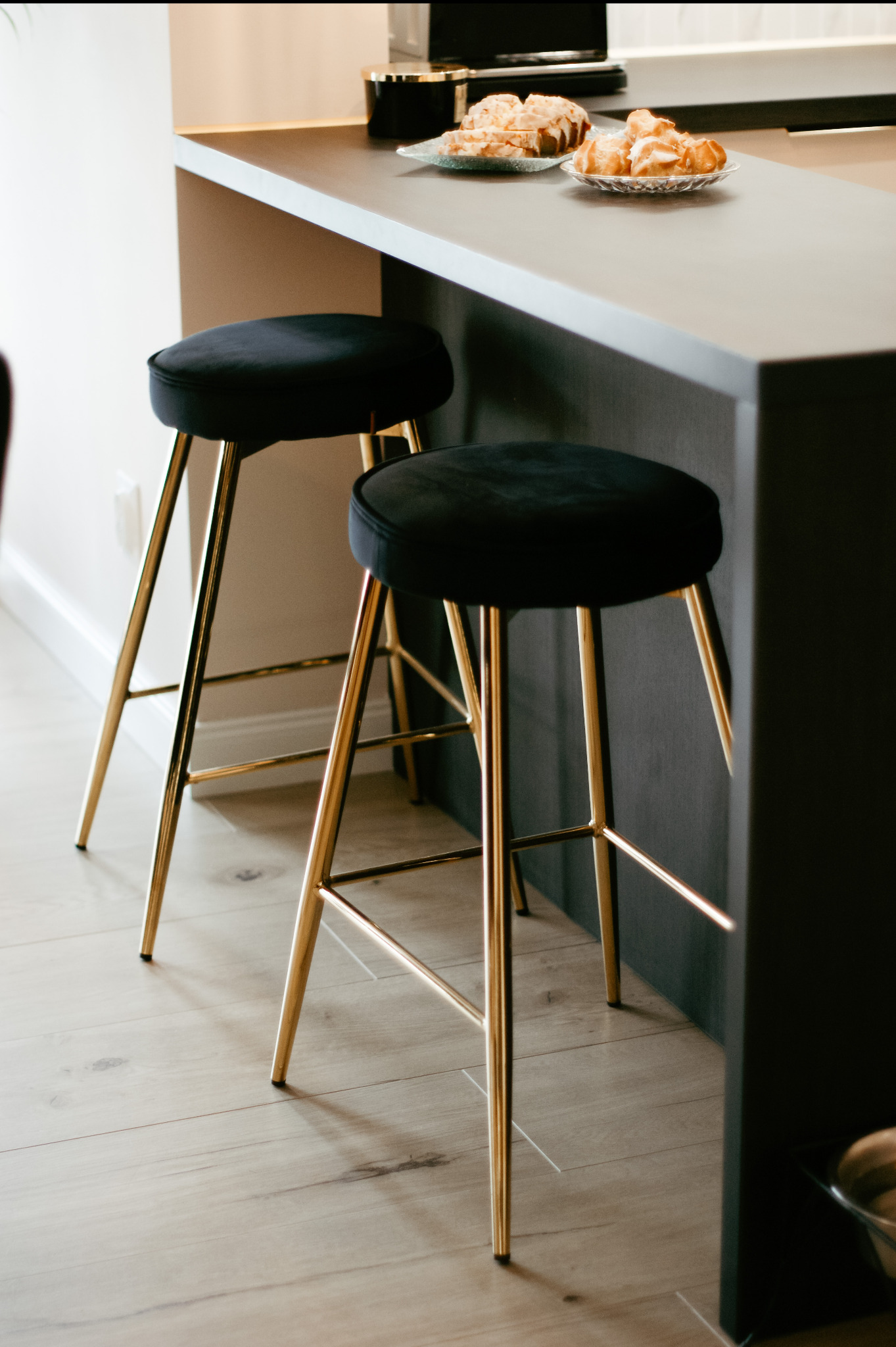 Dwa welurowe hokery z pozłacanymi nogami przy ciemnoszarym blacie kuchennym, na którym leżą ciastka na szklanych talerzykach.