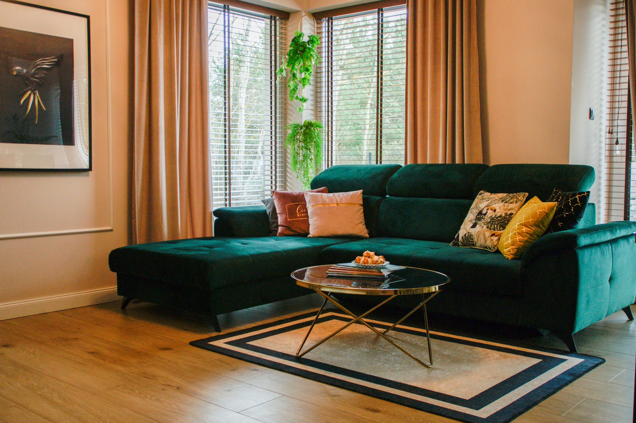 Salon z zielonym narożnikiem, okrągłym stolikiem kawowym na metalowych nóżkach, drewnianą podłogą i zasłonami w odcieniach beżu. Na ścianie wisi obraz w czarnej ramie.