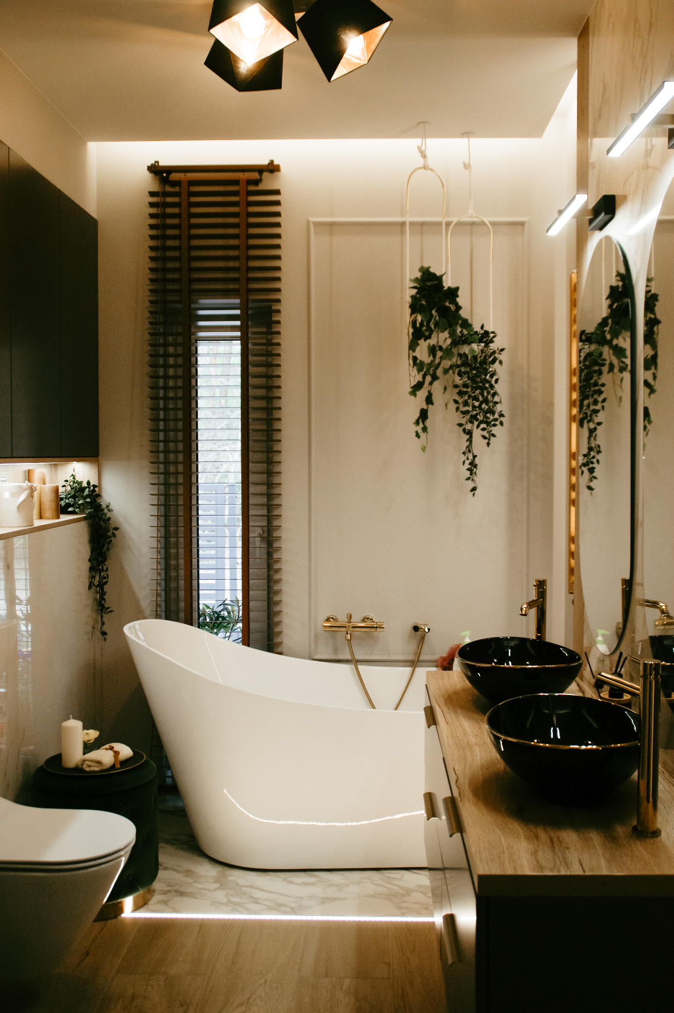 Nowoczesna łazienka z wolnostojącą białą wanną, czarnymi umywalkami nablatowymi ze złotymi akcentami, oknem z drewnianymi żaluzjami i dekoracyjnymi roślinami zwisającymi z sufitu.