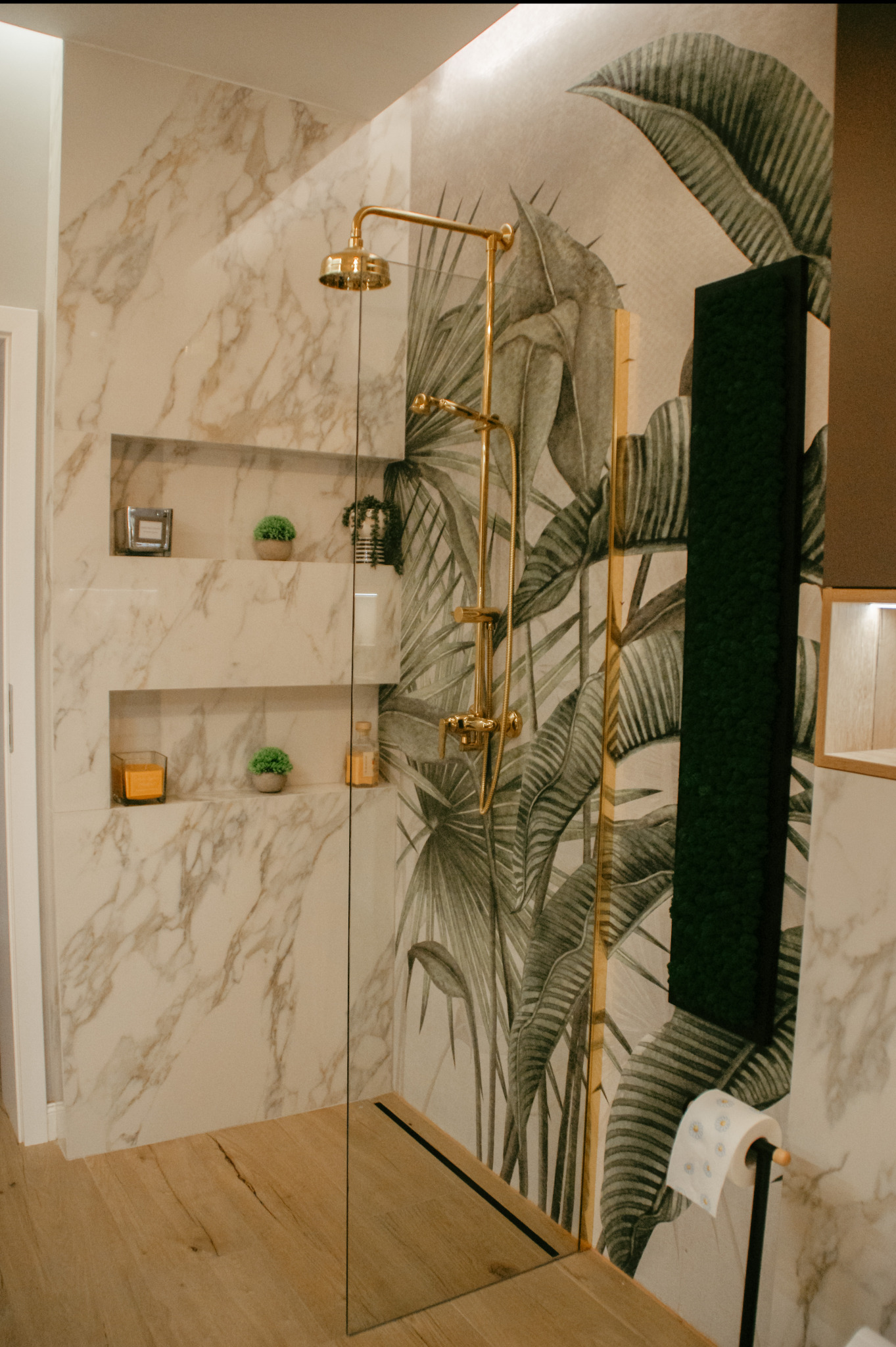 Nowoczesna kabina prysznicowa walk-in z marmurowymi ścianami, złotą armaturą i motywem tropikalnych liści na ścianie, uzupełniona o półki wnękowe z dekoracjami i drewnianą podłogę.