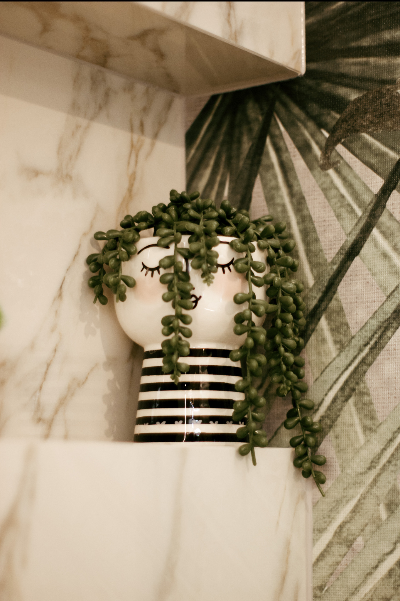 Dekoracyjny ceramiczny wazon w biało-czarne paski z namalowaną twarzą i zwisającą zieloną rośliną, umieszczony na marmurowej półce na tle tapety w liście palmowe.