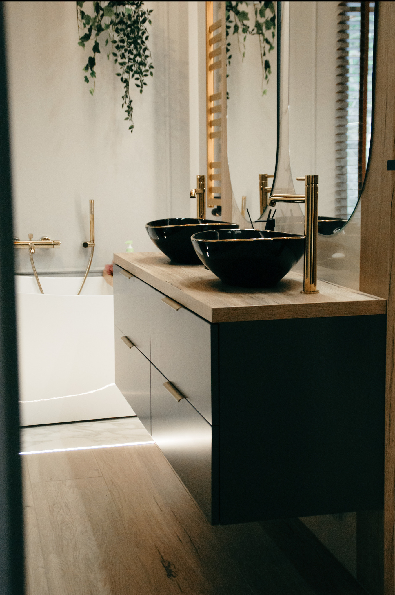 Nowoczesna łazienka z dwoma czarnymi umywalkami na drewnianym blacie, złotą armaturą, podświetleniem LED przy podłodze i dekoracyjną roślinnością.