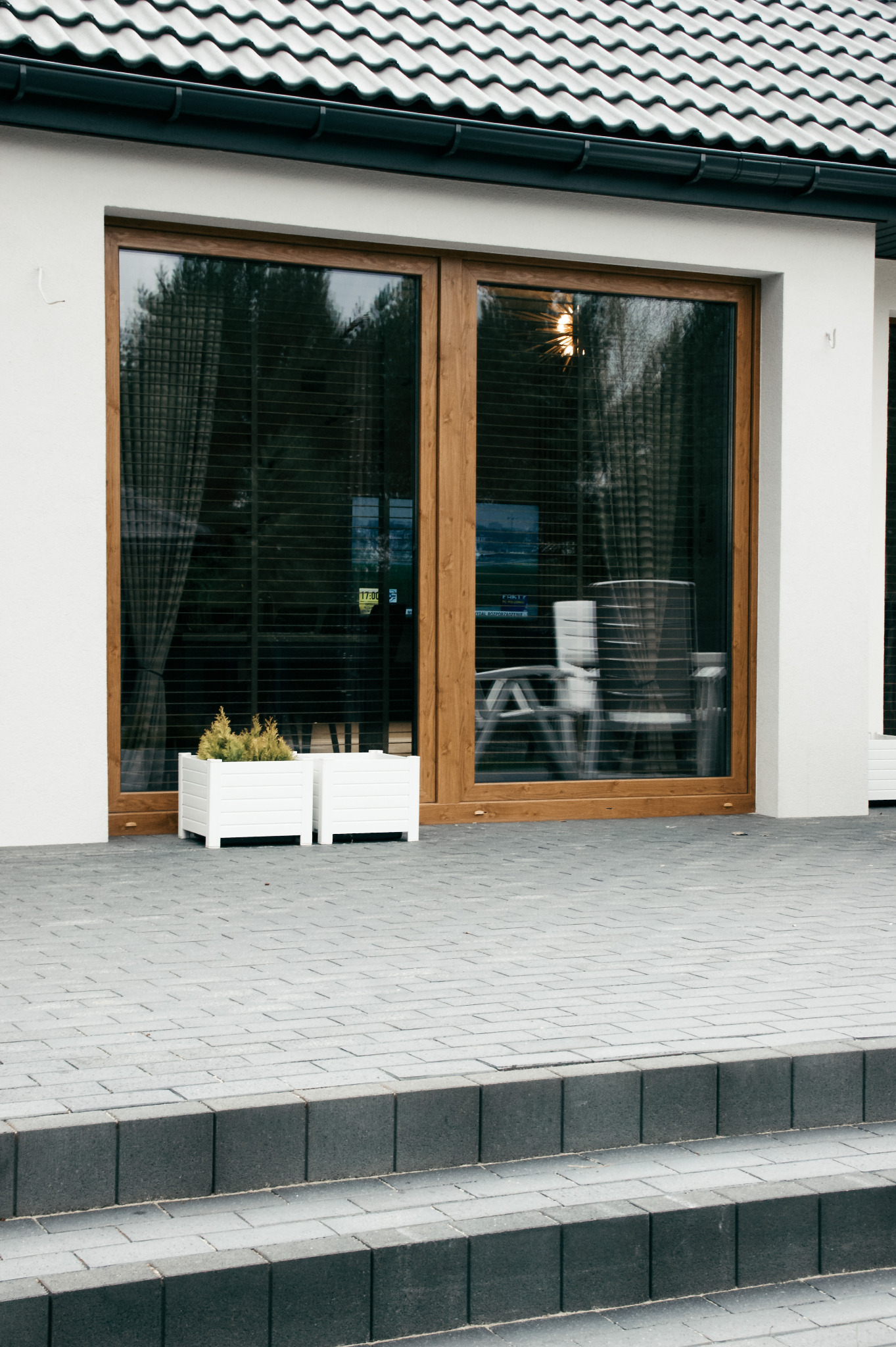 Nowoczesny dom z dużymi przesuwnymi drzwiami tarasowymi w drewnianej ramie, dwa białe kwietniki z iglakami na zewnątrz, szara kostka brukowa na tarasie i schodach.