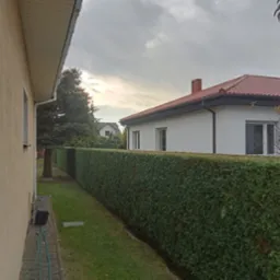 Długi, równo przycięty żywopłot z tui szmaragdowej oddzielający posesje, widok z boku domu, w tle biały dom z czerwoną dachówką i pochmurne niebo.