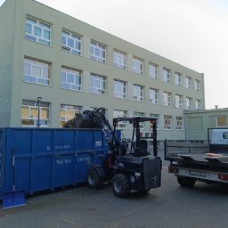 MARIO SUROWCE Mariusz Wojciechowski - Czarny miniładowarka Wolf 750 z pełną łyżką liści i gałęzi wyładowuje je do niebieskiego kontenera SAN-EKO na tle budynku szkoły. Obok stoi biała ciężarówka z opuszczoną platformą i grabiami.