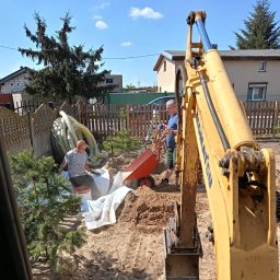 MARIO SUROWCE Mariusz Wojciechowski - Prace ziemne w ogrodzie: żółta koparka w trakcie wykopywania ziemi, obok pracownik siedzący na wiadrze i układający białą tkaninę, drugi pracownik z niwelatorem i taczką w tle.