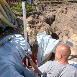 MARIO SUROWCE Mariusz Wojciechowski - Pracownik w szarej koszulce umieszcza rurę drenarską w wykopie, widoczny niwelator optyczny, poziomica i rury drenażowe w tle, folia ochronna rozłożona w wykopie.