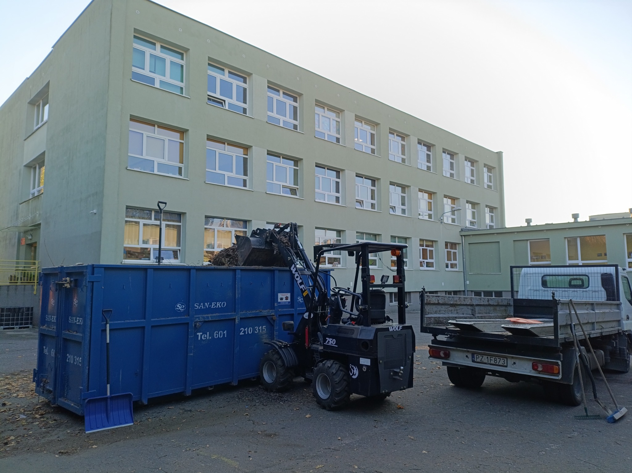 Czarny miniładowarka Wolf 750 z pełną łyżką liści i gałęzi wyładowuje je do niebieskiego kontenera SAN-EKO na tle budynku szkoły. Obok stoi biała ciężarówka z opuszczoną platformą i grabiami.