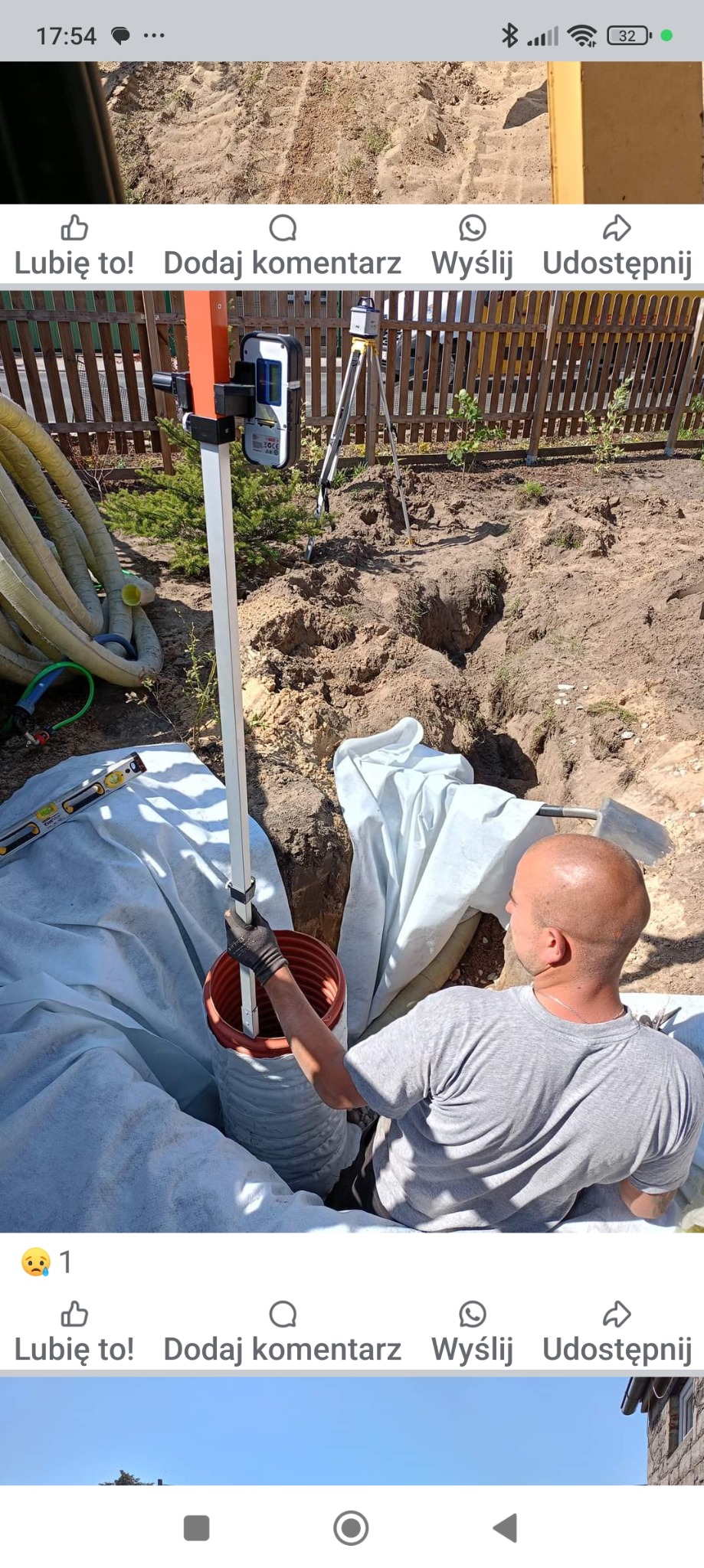 Pracownik w szarej koszulce umieszcza rurę drenarską w wykopie, widoczny niwelator optyczny, poziomica i rury drenażowe w tle, folia ochronna rozłożona w wykopie.