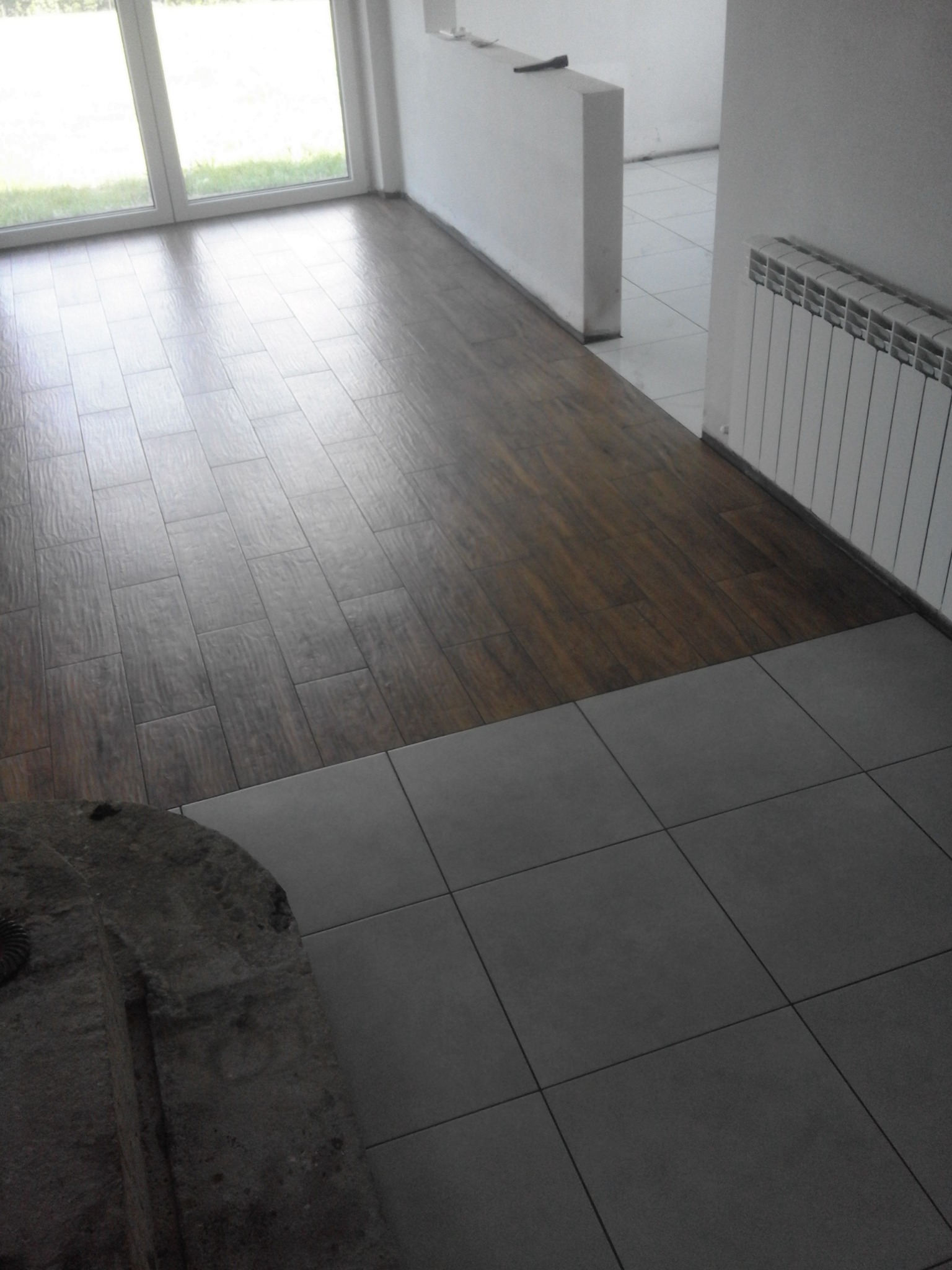 Fragment wnętrza po remoncie z widocznym połączeniem ciemnych paneli podłogowych imitujących drewno i szarych płytek ceramicznych, grzejnik na ścianie.