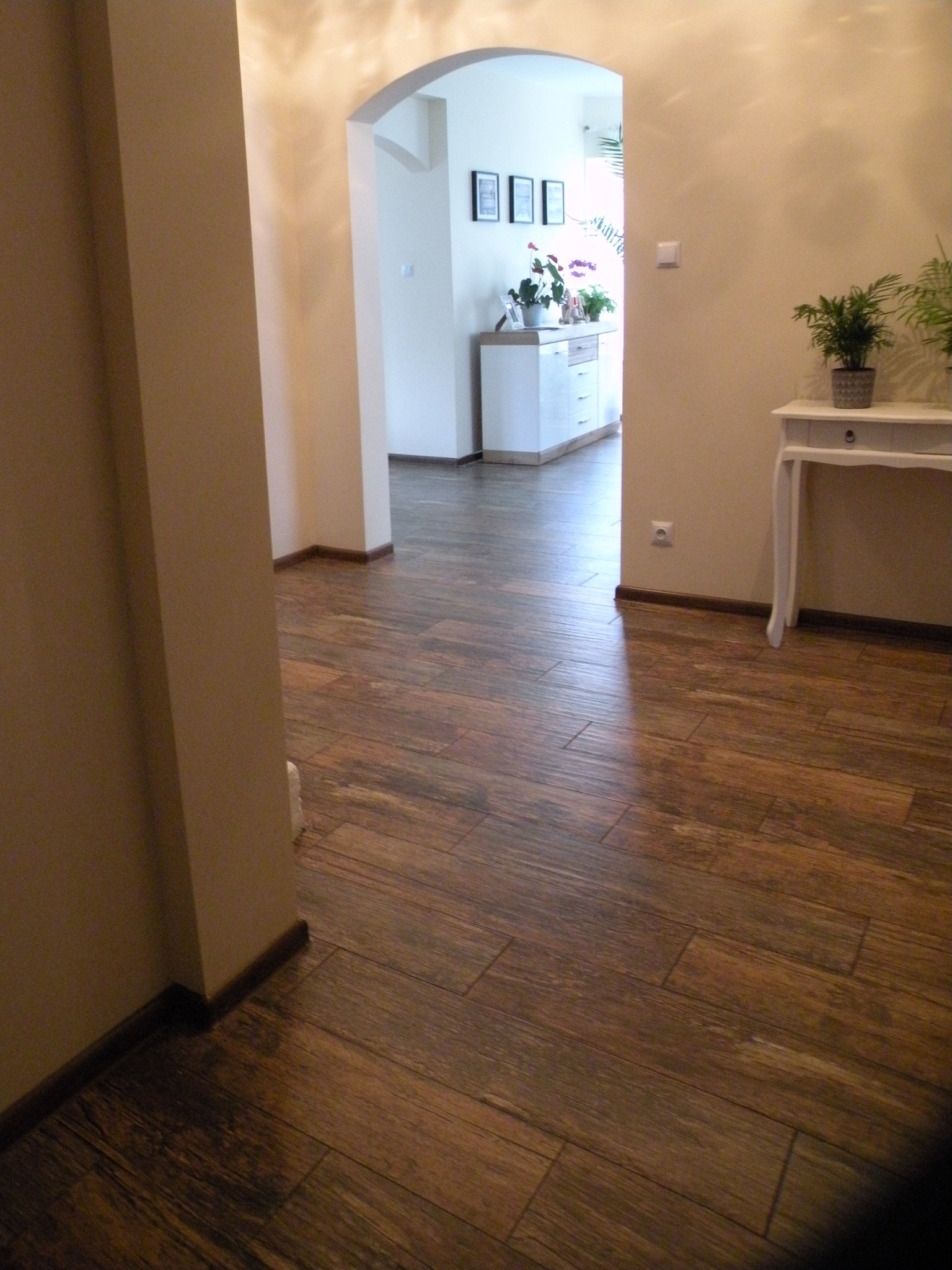 Ułożone panele podłogowe w odcieniach brązu, imitujące drewno, w korytarzu z łukowatym przejściem do jasnego pomieszczenia z białymi meblami i roślinami.