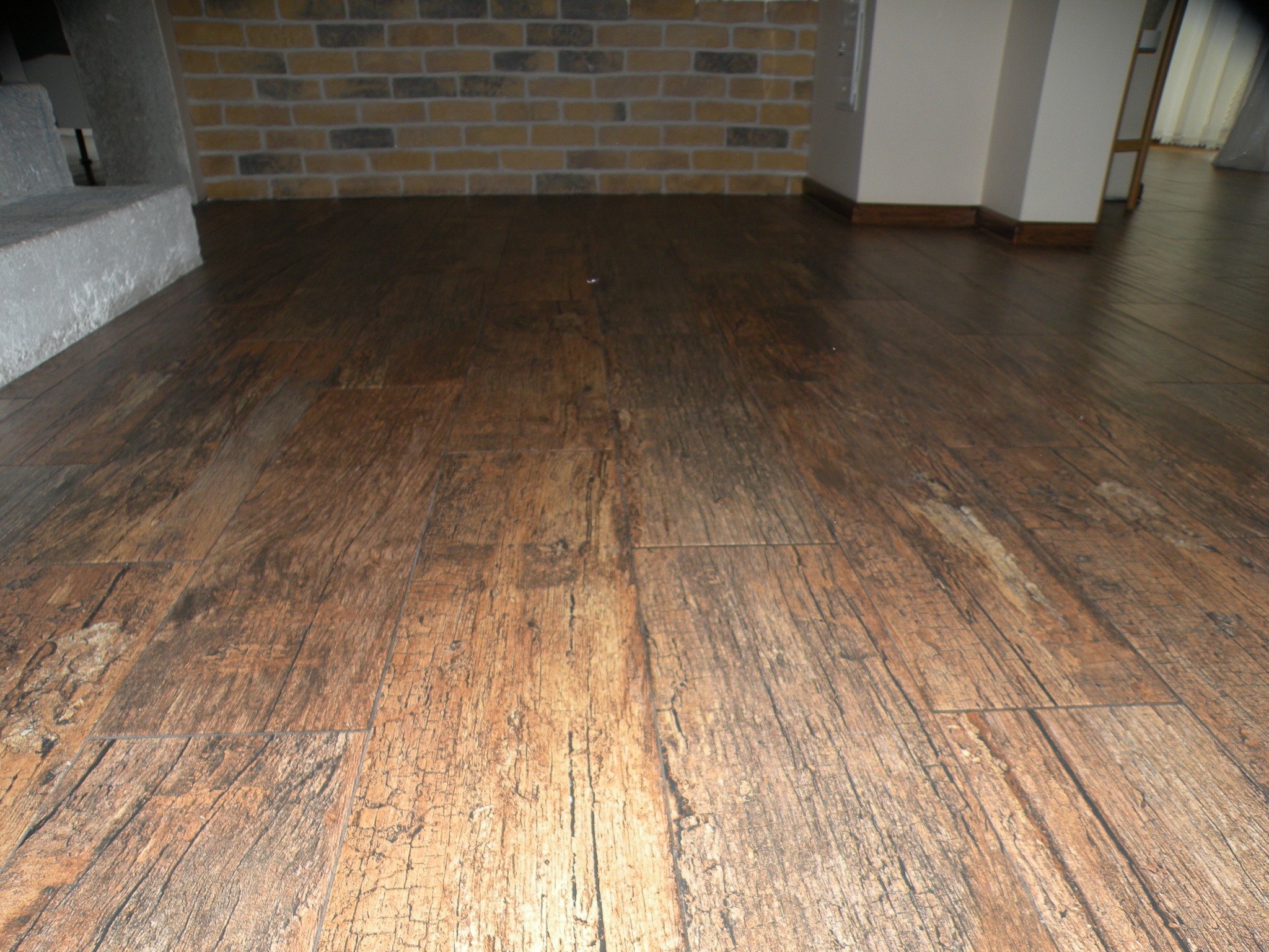 Ułożone panele podłogowe imitujące ciemne drewno z wyraźną fakturą słojów, widoczne fragmenty ściany z cegły oraz schodów.