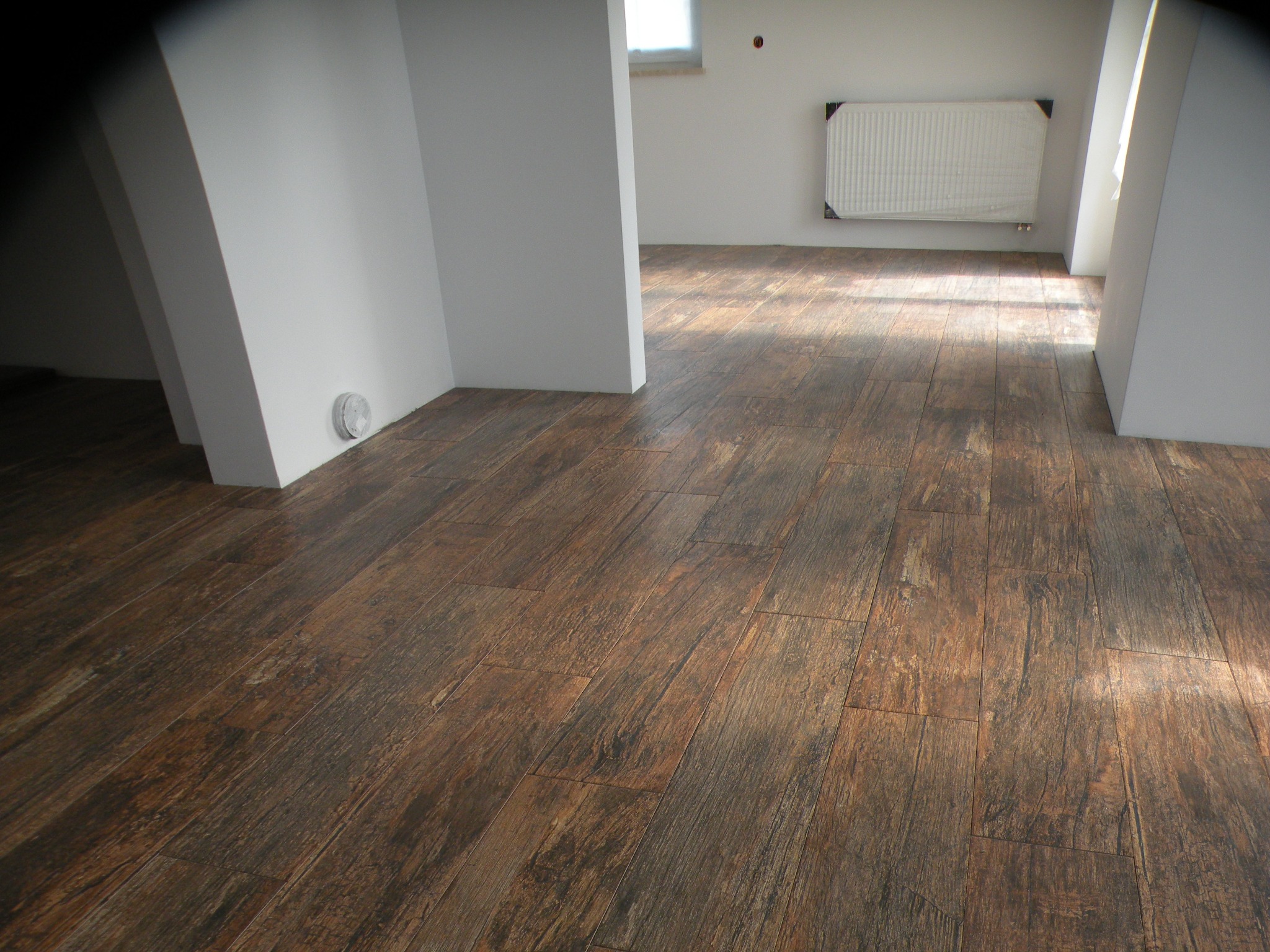 Ułożona podłoga z paneli imitujących ciemne drewno, widok na fragment pomieszczenia z białymi ścianami i grzejnikiem, światło słoneczne podkreśla strukturę paneli.