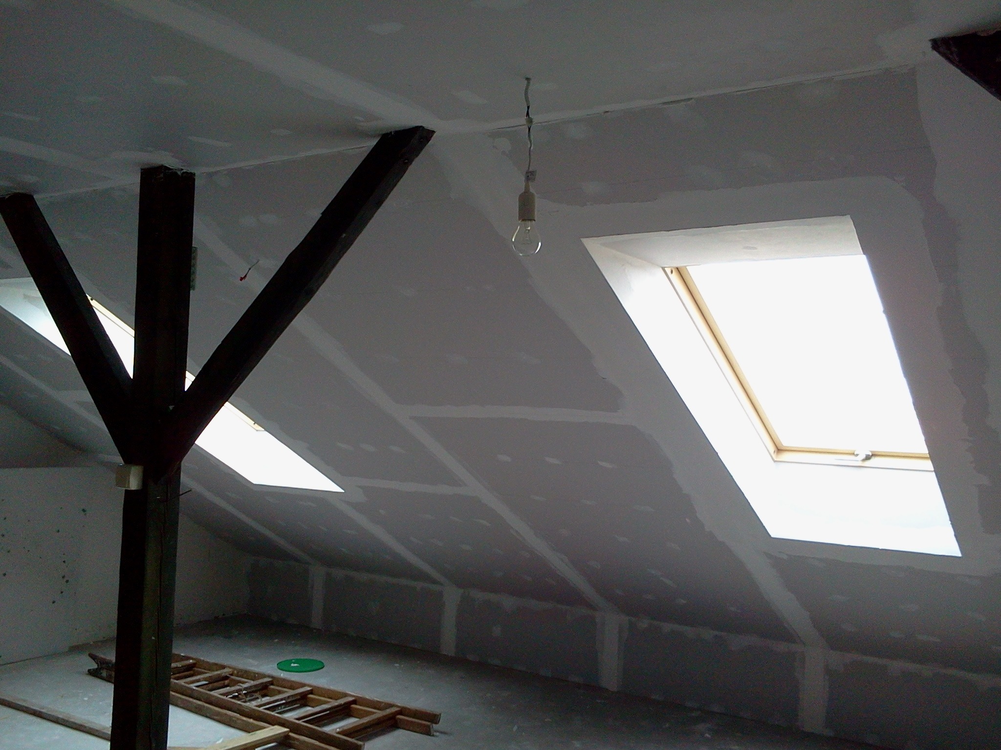 Poddasze w trakcie prac wykończeniowych: zamontowane płyty gipsowo-kartonowe, widoczne drewniane elementy konstrukcyjne, okna dachowe i prowizoryczne oświetlenie.