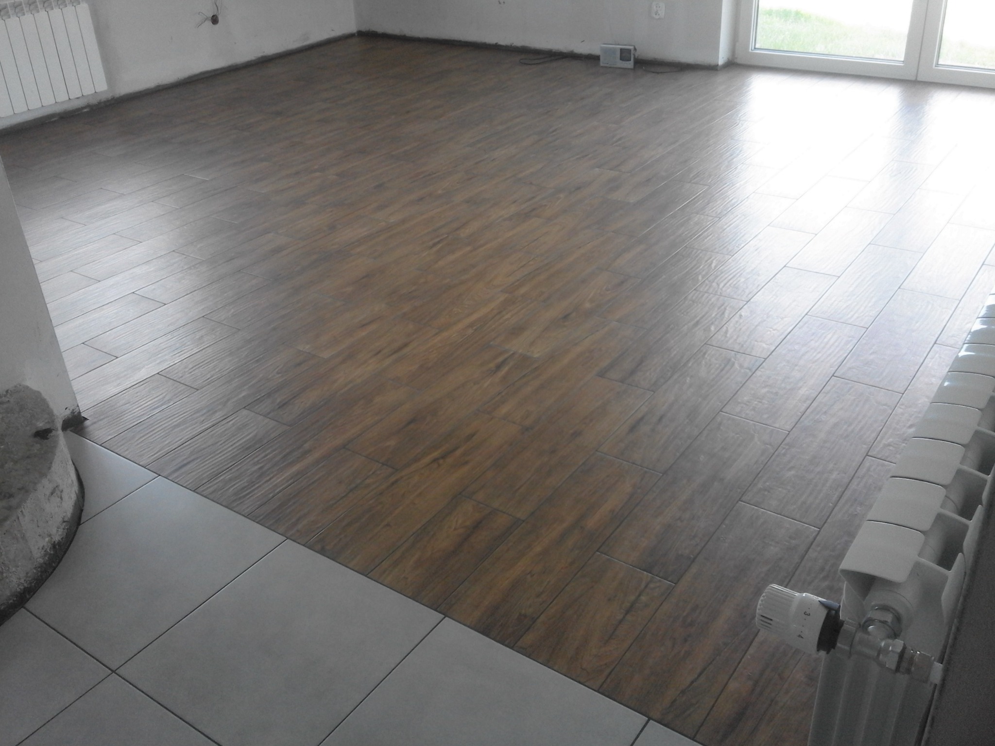 Ułożona podłoga z paneli imitujących drewno w jasnym pomieszczeniu, widoczny fragment grzejnika i szare płytki podłogowe przy kolumnie.