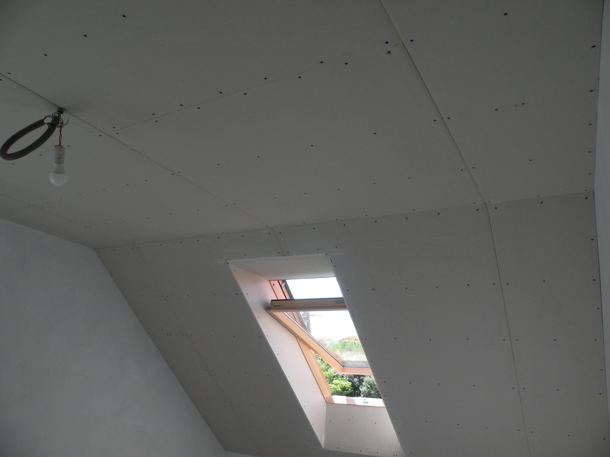 Poddasze w trakcie wykańczania: widoczne płyty gipsowo-kartonowe na skośnym suficie, okno dachowe z drewnianą ramą oraz tymczasowe oświetlenie w postaci żarówki na kablu.