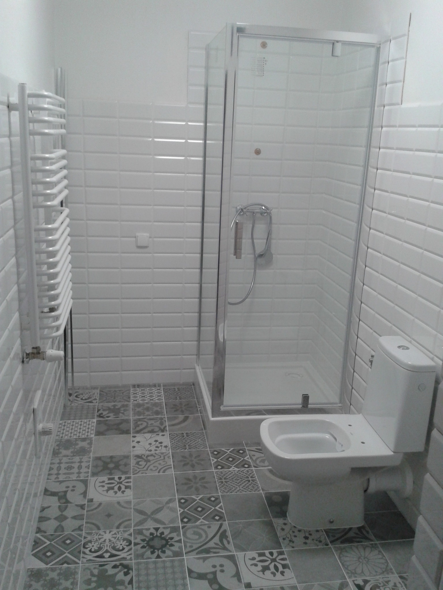 Wykończona łazienka z białą armaturą, kabiną prysznicową i dekoracyjnymi płytkami podłogowymi w odcieniach szarości i bieli, ściany wyłożone białymi, podłużnymi płytkami.