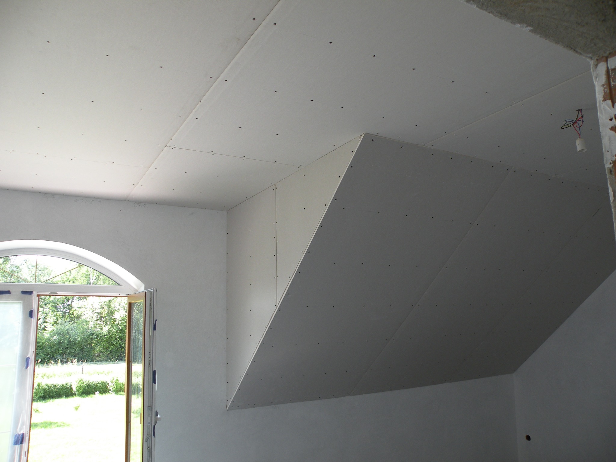 Poddasze w trakcie izolacji w Warszawie, widoczne płyty gipsowo-kartonowe zamontowane na skośnej powierzchni, okno z widokiem na zieleń.