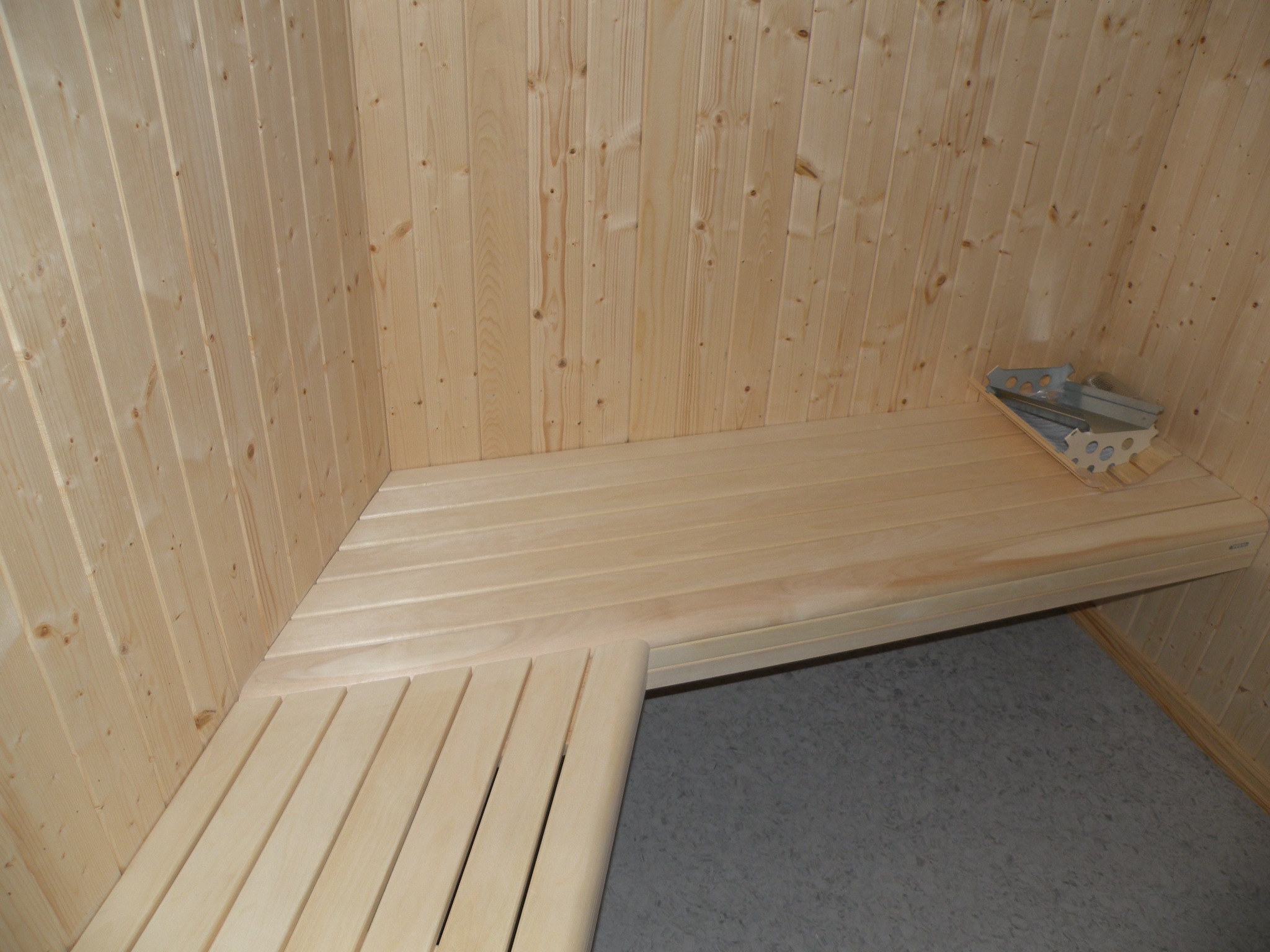 Wnętrze sauny z jasnego drewna, z ławkami na różnych poziomach i metalowym koszem na kamienie.