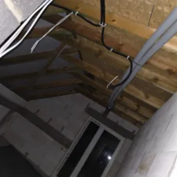 Wnętrze poddasza w trakcie prac: widoczne kable elektryczne przymocowane do drewnianej konstrukcji dachu, szare rury instalacyjne i surowa ściana z oknem.