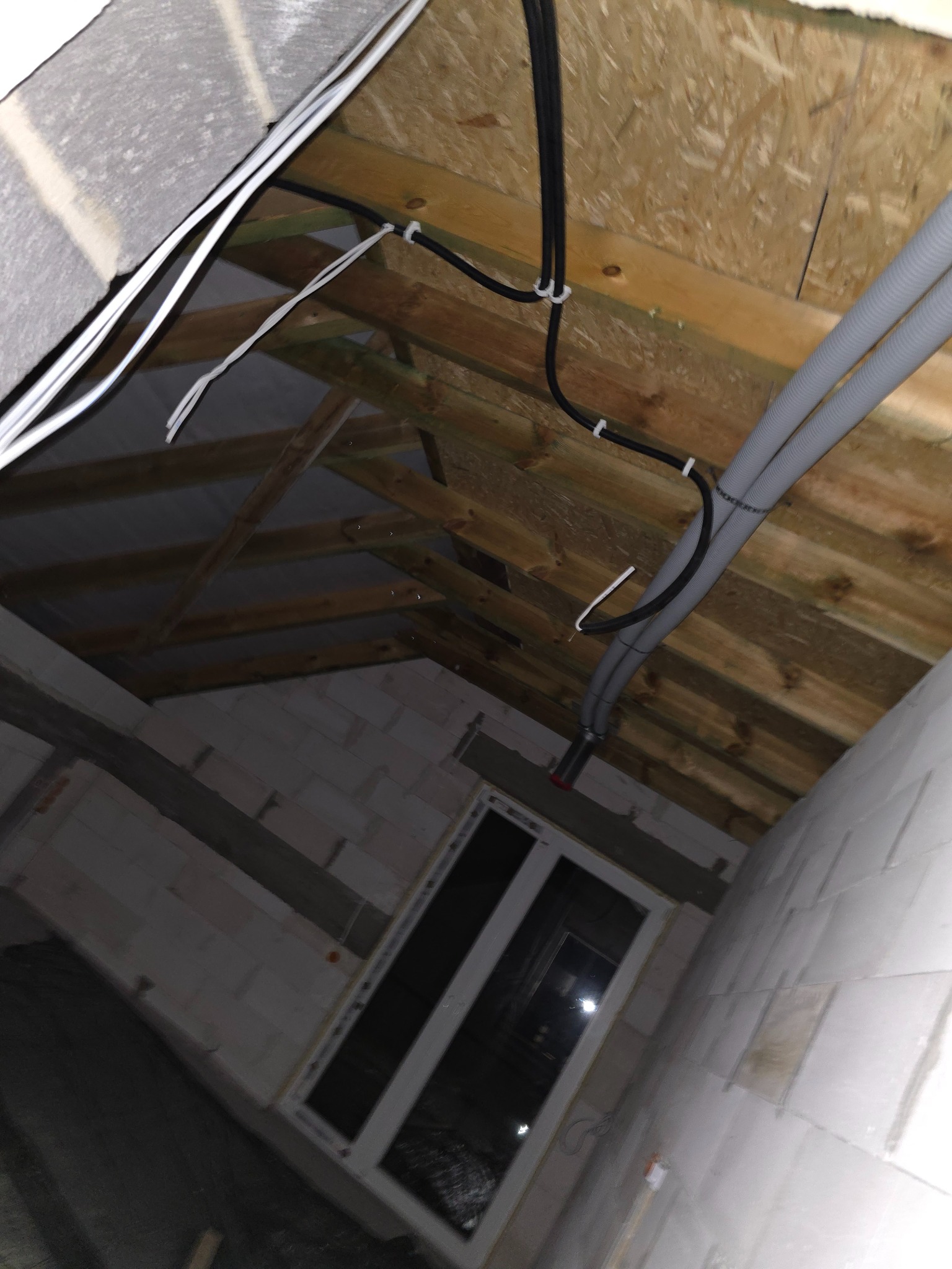 Wnętrze poddasza w trakcie prac: widoczne kable elektryczne przymocowane do drewnianej konstrukcji dachu, szare rury instalacyjne i surowa ściana z oknem.