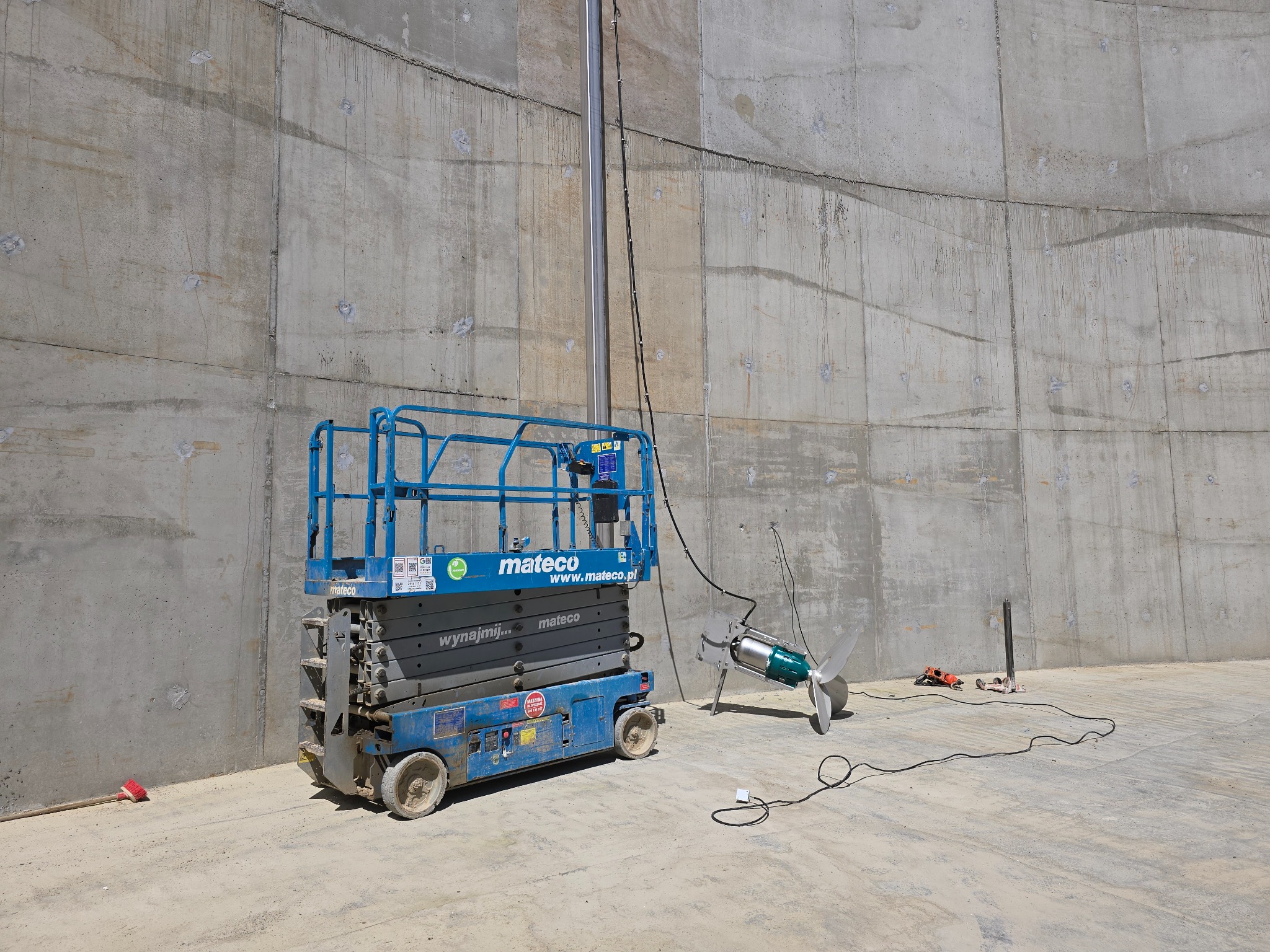 Wnętrze betonowego zbiornika z podnośnikiem Mateco i elementem instalacji z napędem śmigłowym. Widoczne przewody i narzędzia, surowe ściany.