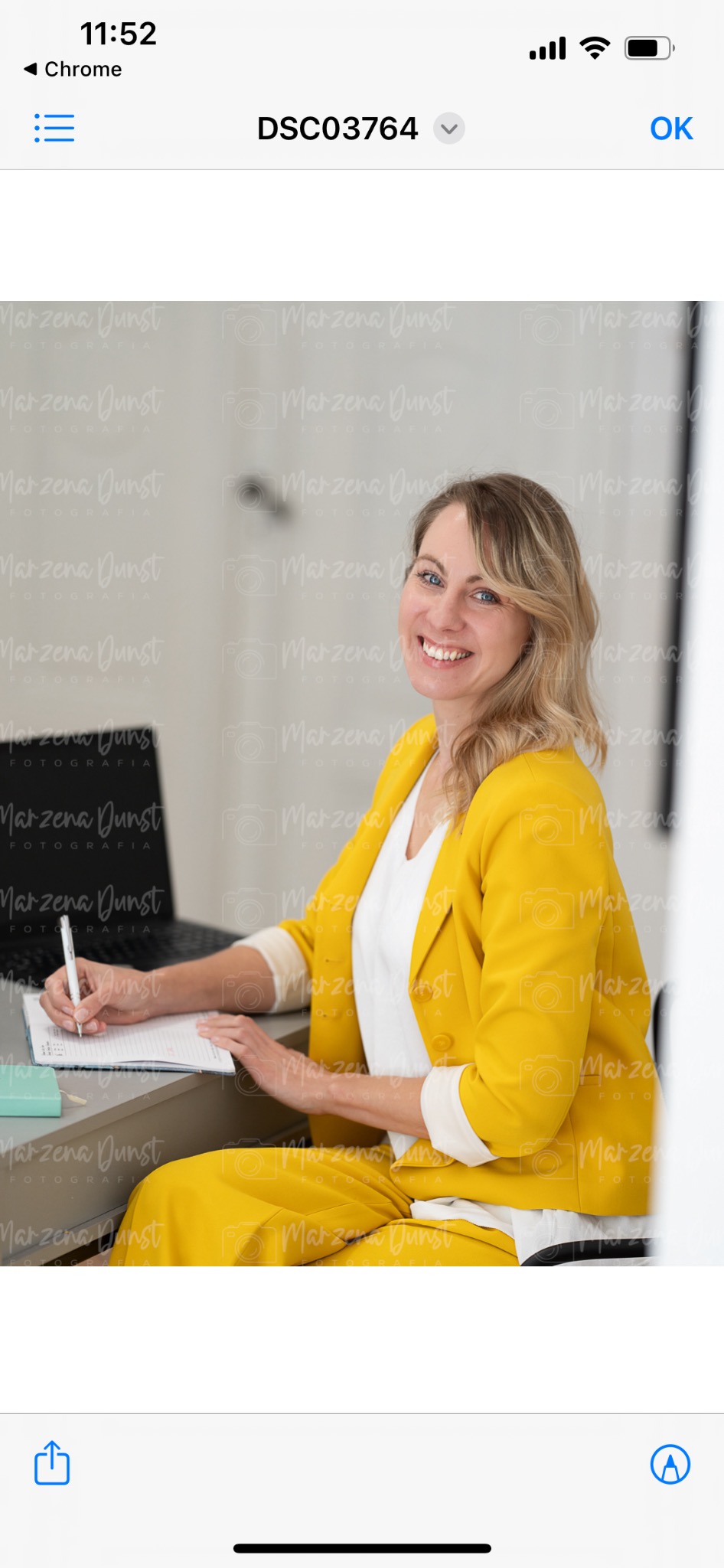 Uśmiechnięta kobieta w żółtym garniturze siedzi przy biurku, pisząc w notatniku białym długopisem, z laptopem w tle i delikatnym tłem z powtarzającym się logo fotografa.